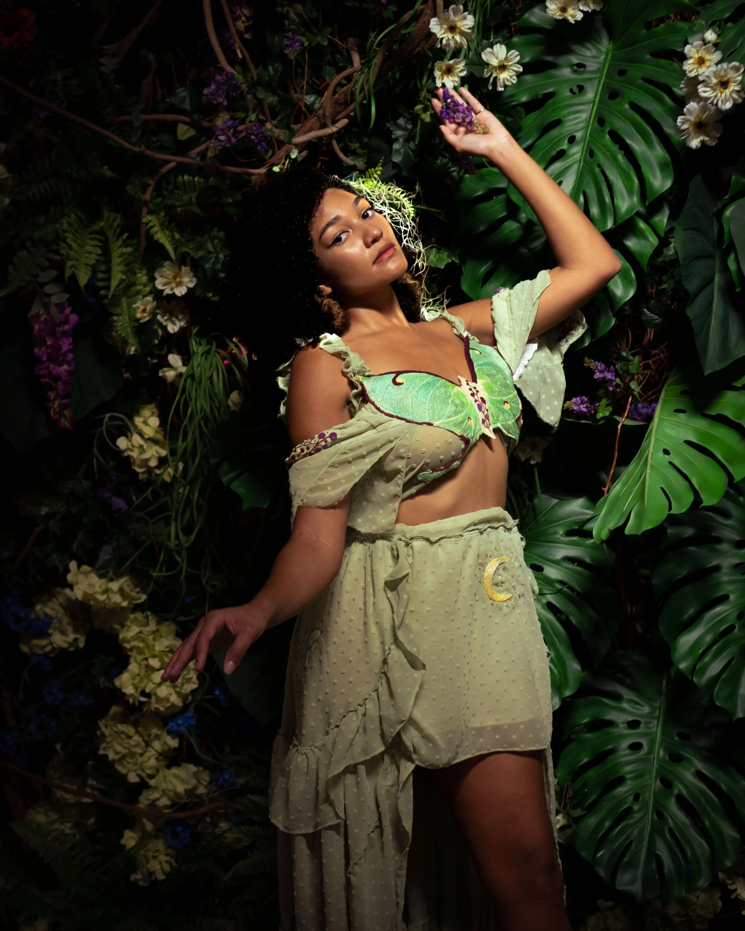 A woman in a flowy, green outfit with butterfly designs, surrounded by lush green leaves and white flowers, posing against a dense foliage background.