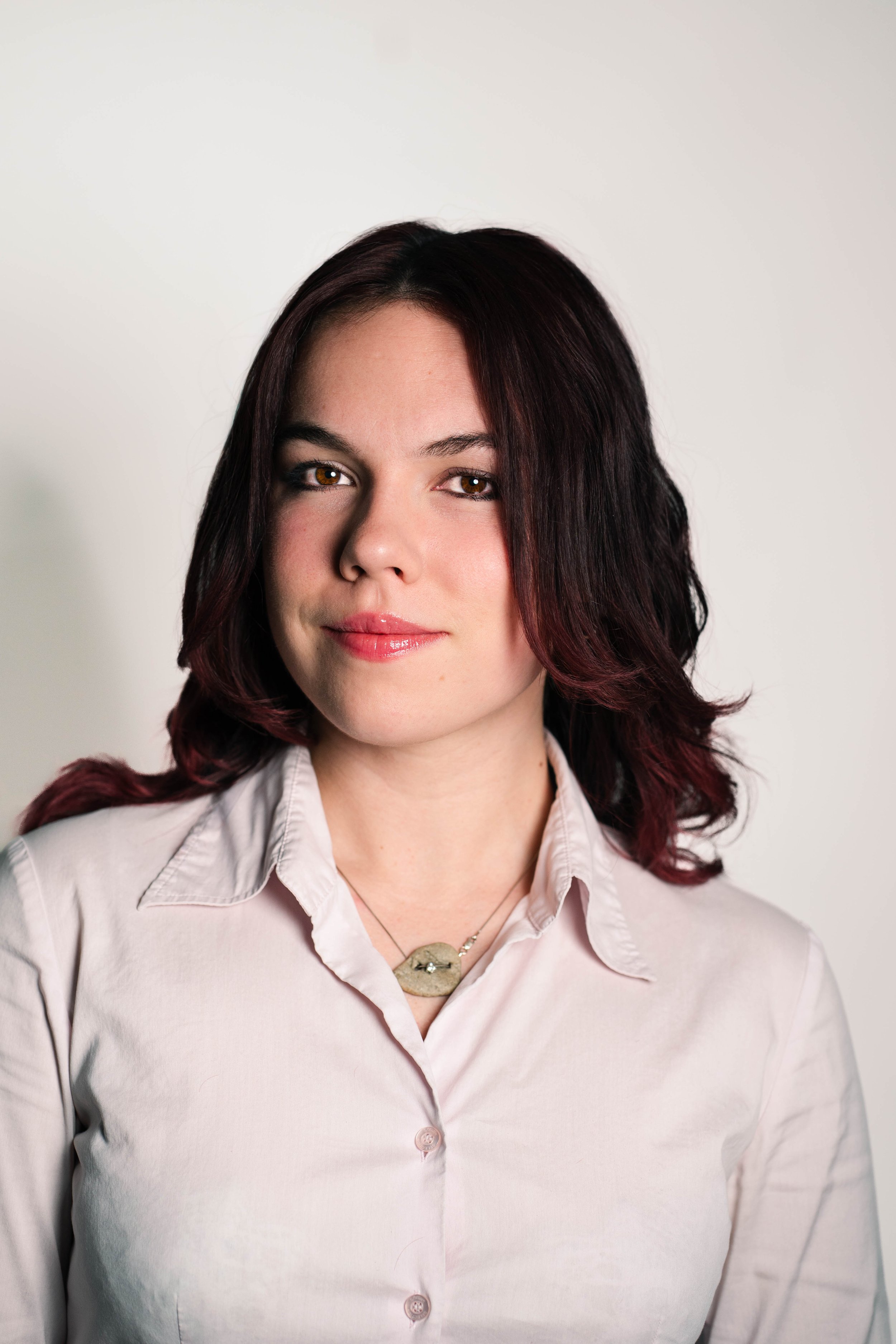 Portrait of a woman with shoulder-length dark hair, wearing a white button-up shirt and a necklace, against a plain light background.