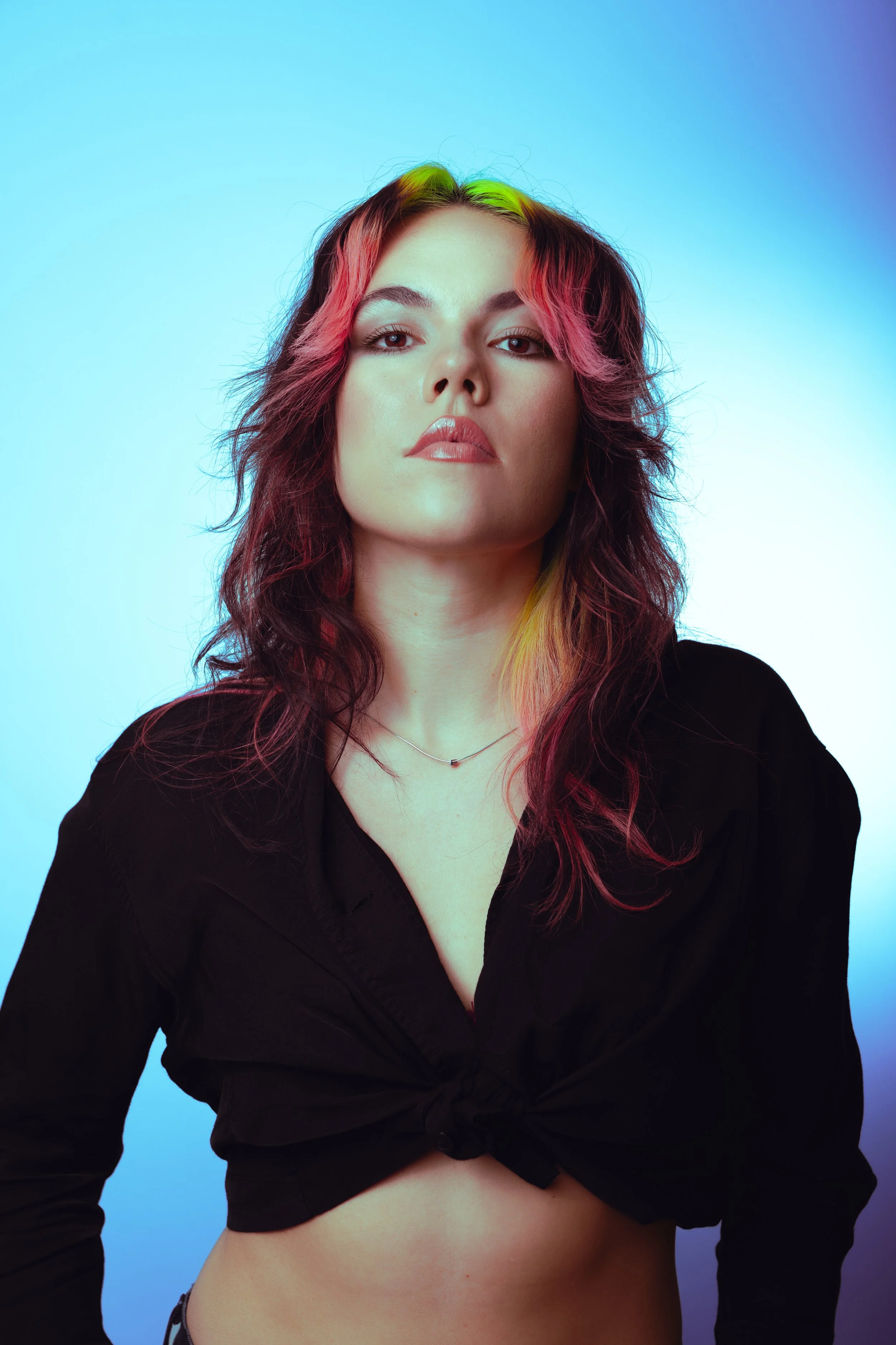 A young woman with multicolored hair, wearing a black tied-up shirt and a delicate necklace, poses against a gradient blue background.