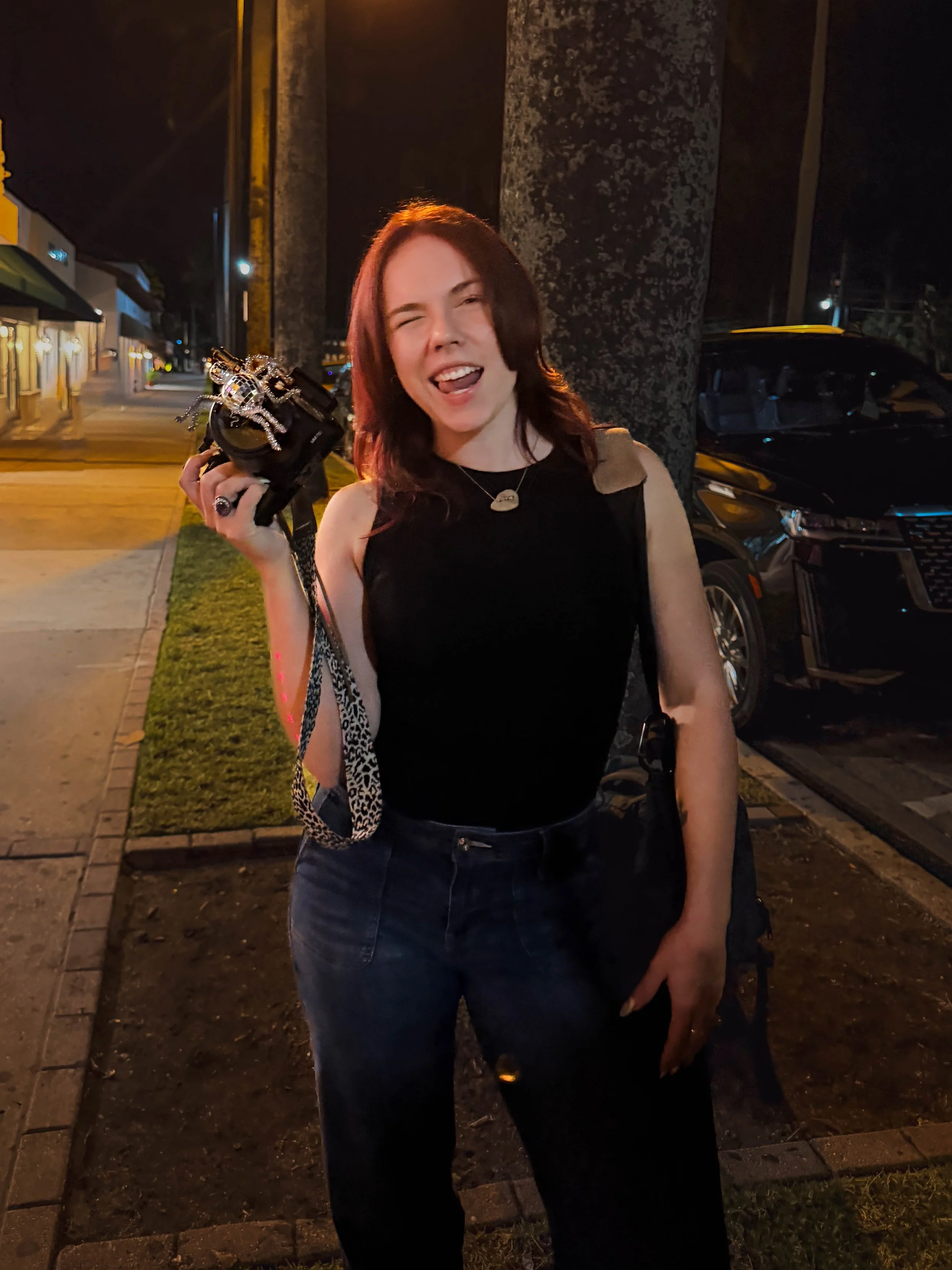 A young woman with red hair making a playful face and sticking her tongue out while holding a camera with a rhinestone decoration in her right hand. She is standing on a sidewalk at night with buildings, cars, and a large stone column in the backgrou