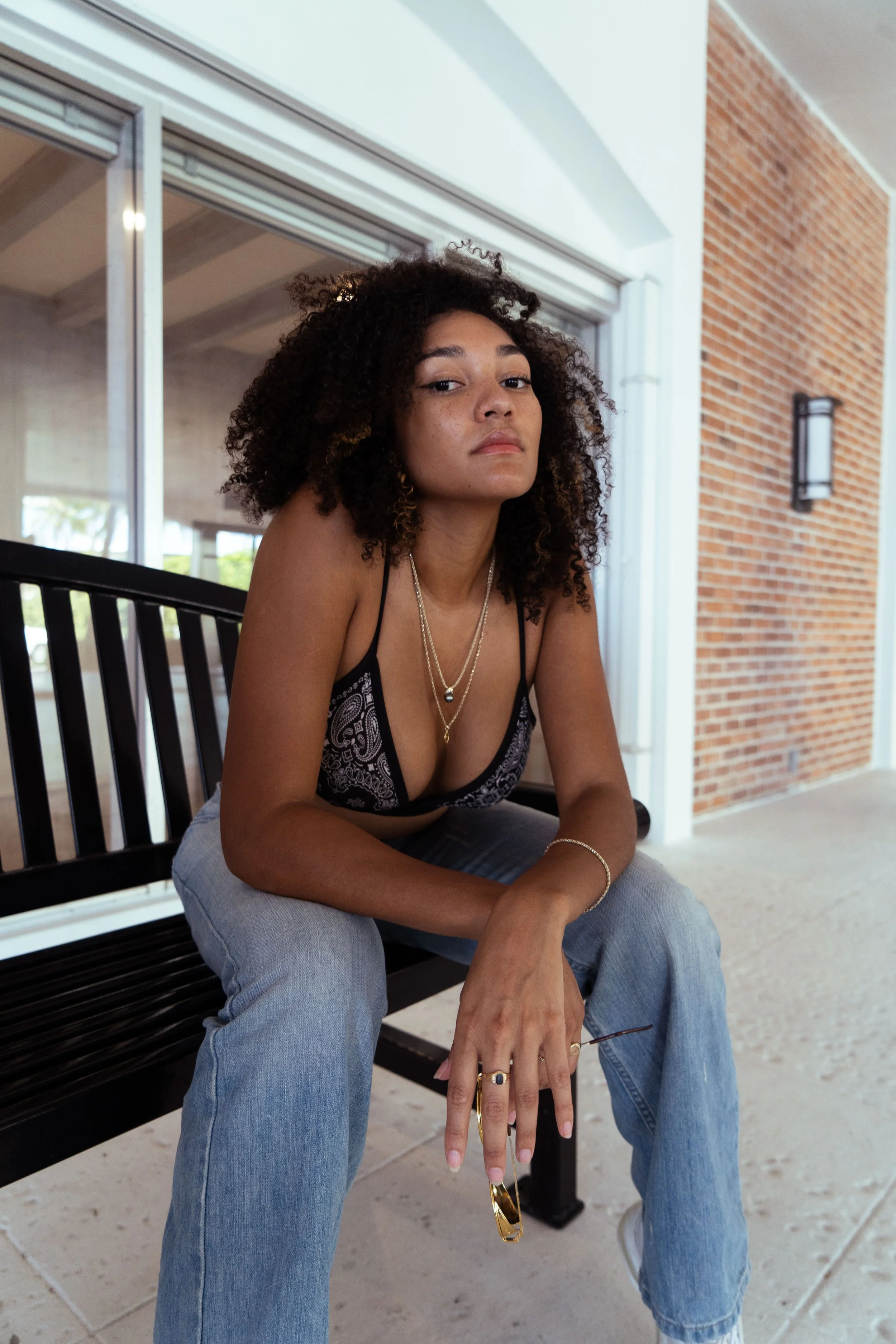 A young woman with curly hair sitting on a black park bench outside, wearing a black tank top with a paisley pattern, light blue jeans, layered necklaces, and sunglasses hanging from her finger, with a brick wall and glass door behind her.