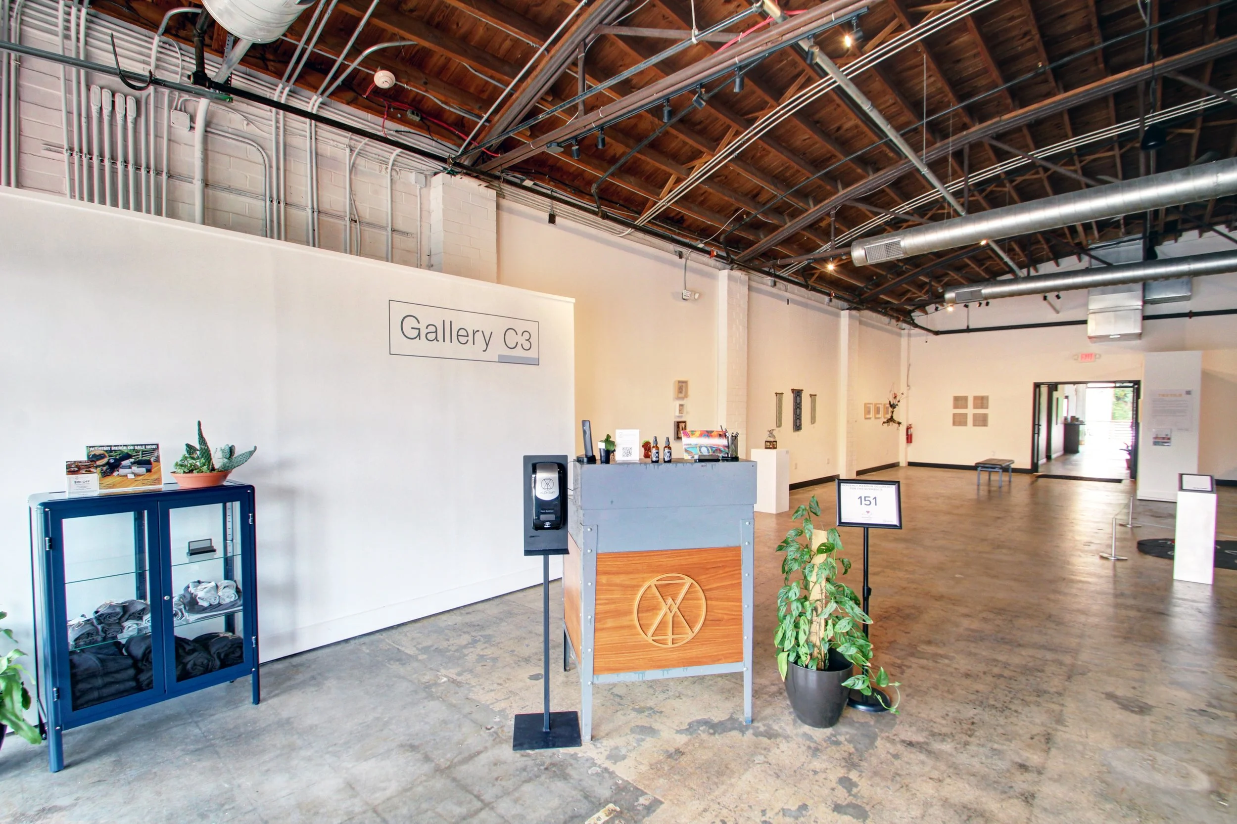 Interior of an art gallery with white walls, wood ceiling, and artwork displayed. A black reception desk, a plant, and a glass display case are visible. Sign reads 'Gallery C3'.
