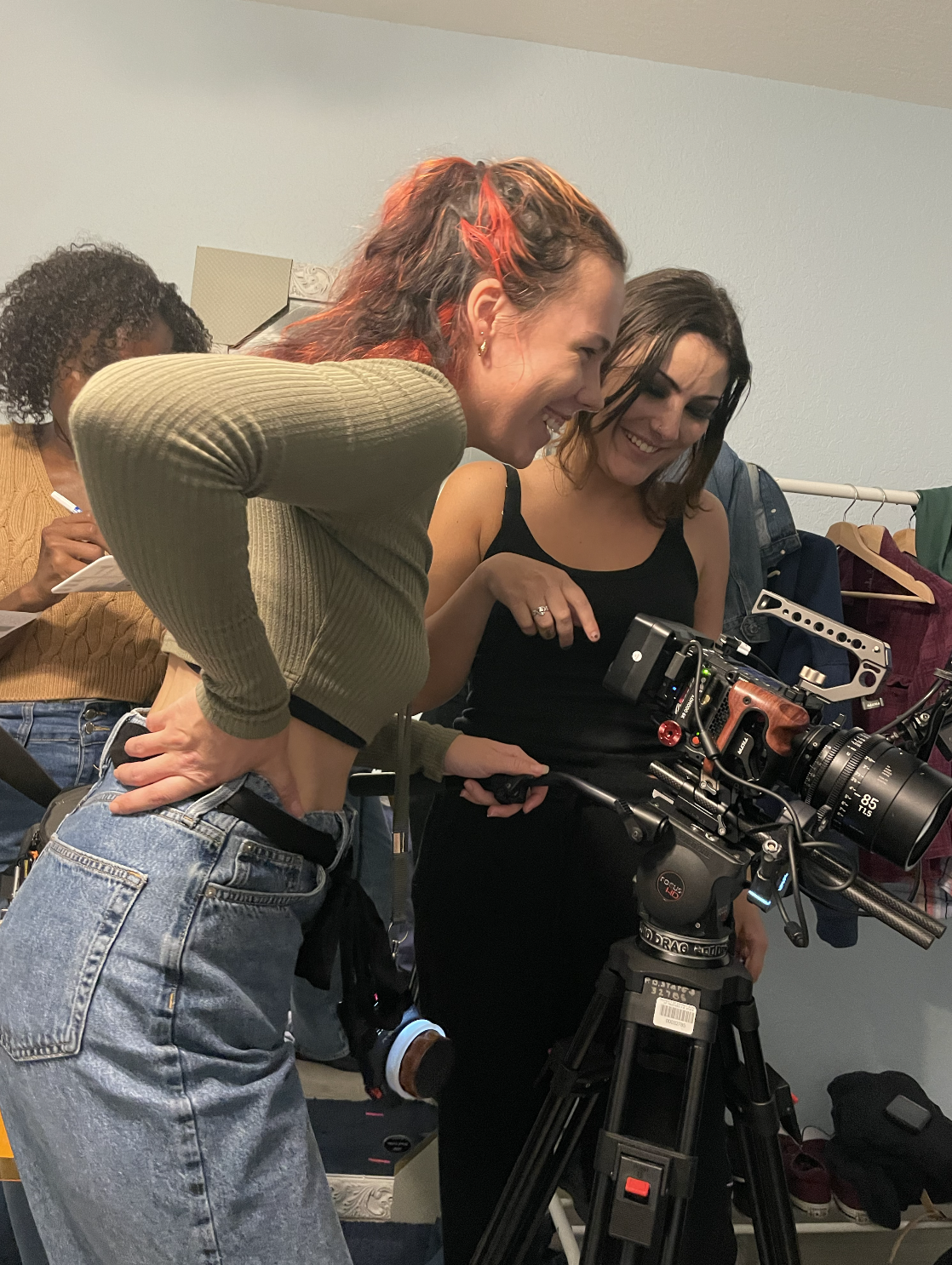 Two women looking at a professional video camera on a tripod, smiling and laughing, on a film set or production indoors.