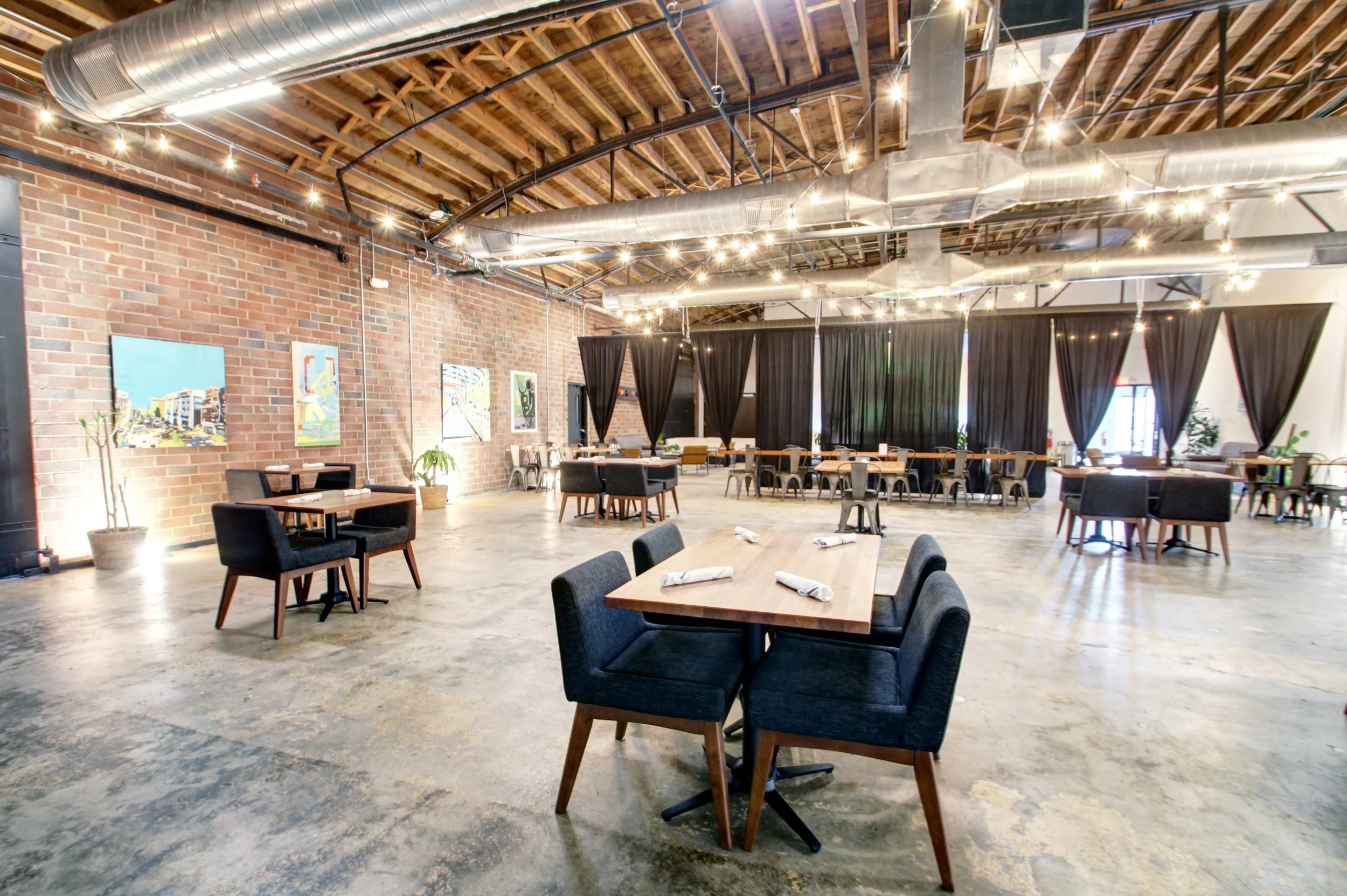 Interior of a modern restaurant with wooden ceiling, brick walls, black curtains, potted plants, and multiple tables set with napkins, with artwork on walls and string lights hanging from the ceiling.