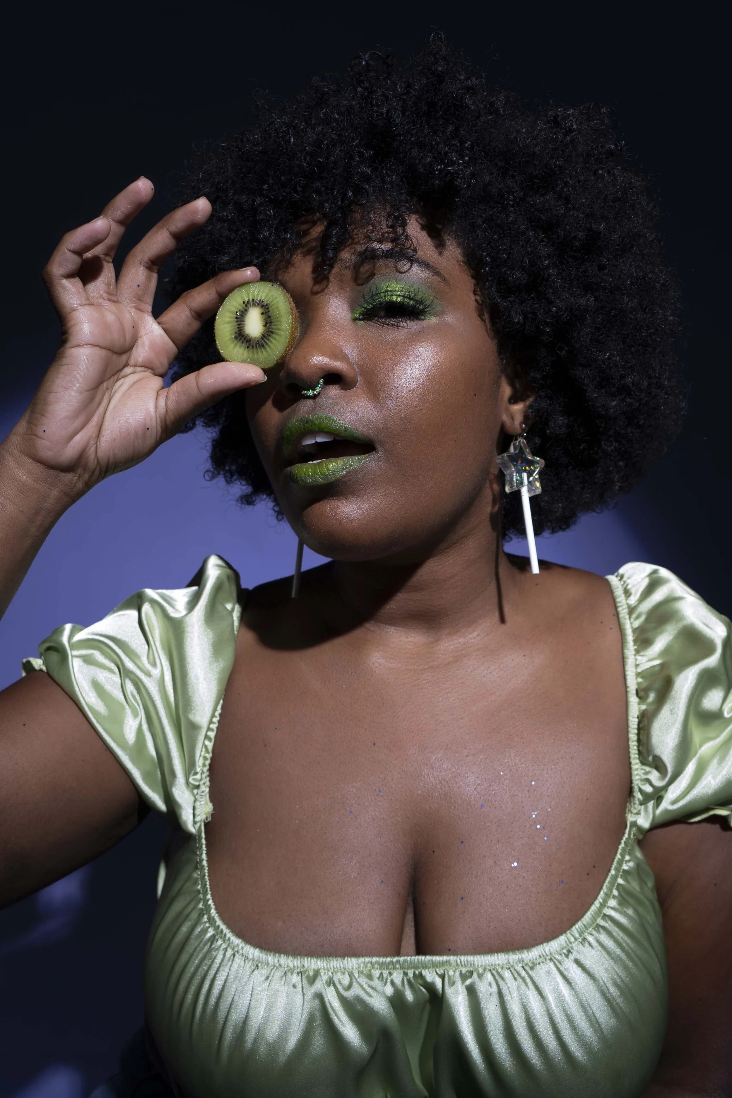 A woman with dark skin and curly black hair holds a slice of kiwi fruit to her right eye. She wears green eye makeup, matching lipstick, and earrings shaped like stars. She is dressed in a shiny, green, puff-sleeve top.