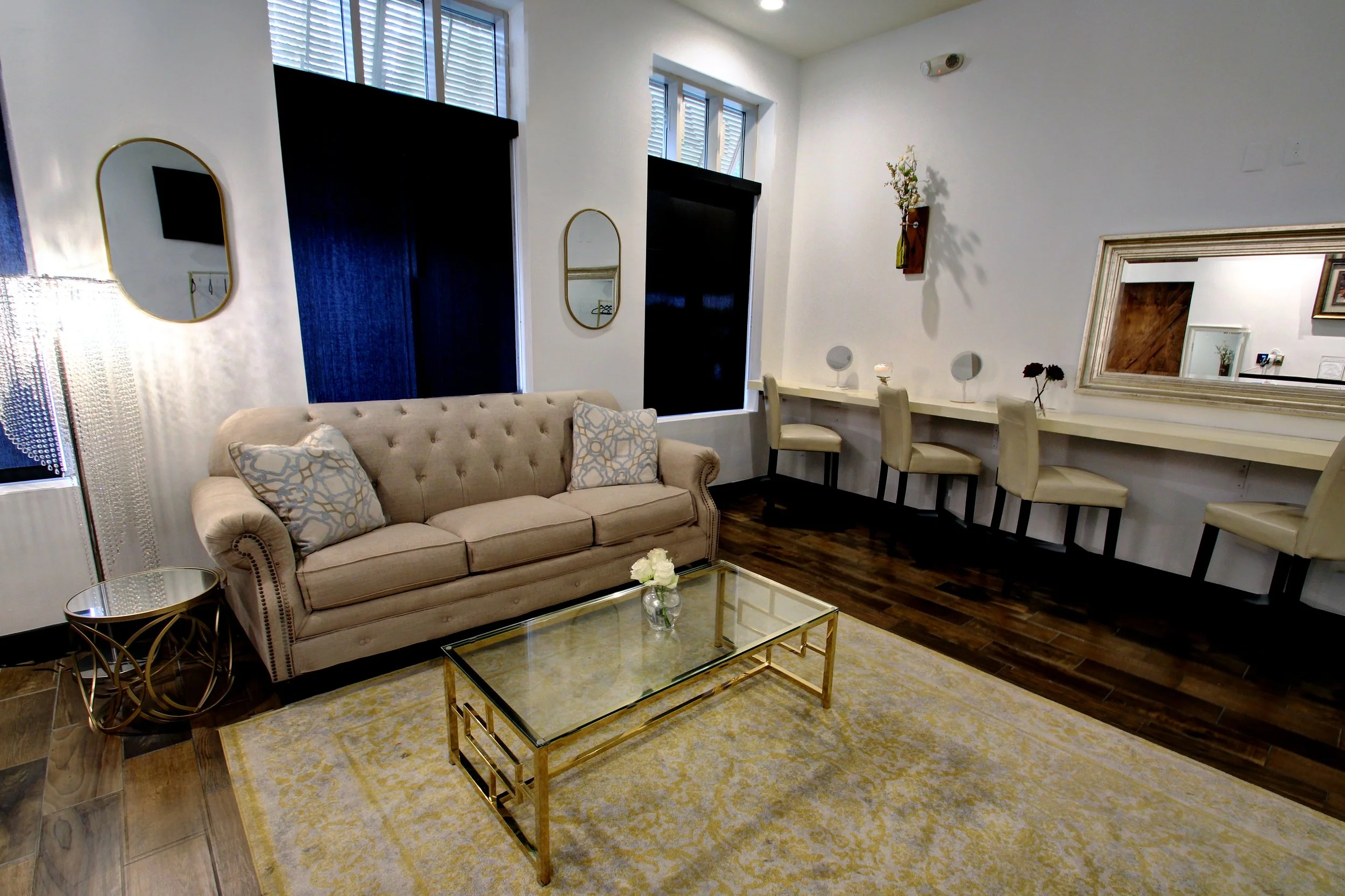A living room with a beige tufted sofa, a glass coffee table with a flower vase, a yellow and white patterned rug, three white bar stools along a counter with a large mirror, decorative mirrors and wall art, and a bright window with blinds.