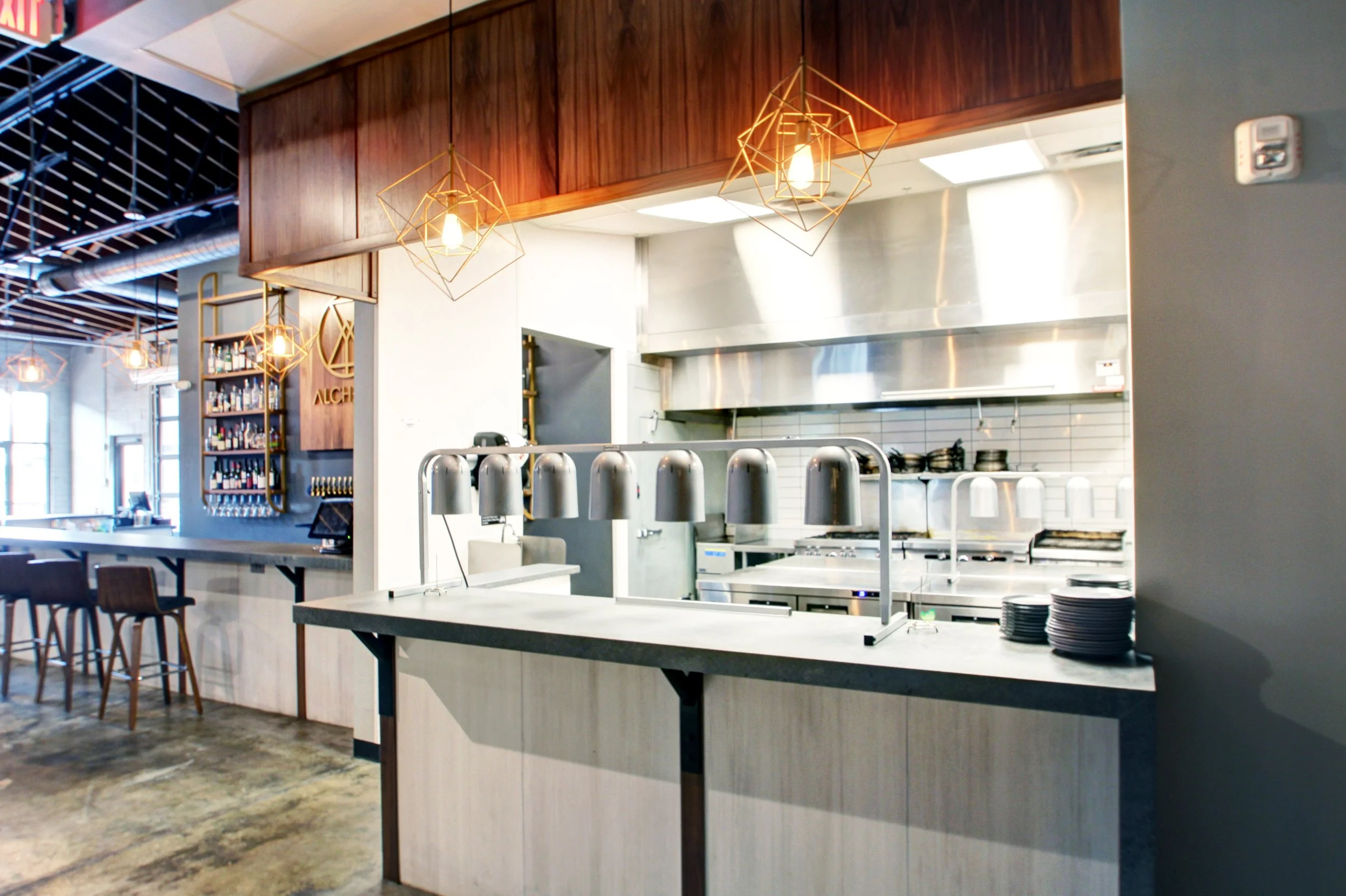 Modern restaurant kitchen with stainless steel appliances, white tile backsplash, and hanging light fixtures over a counter with plates and food warmers, dining area with chairs and tables visible to the left.