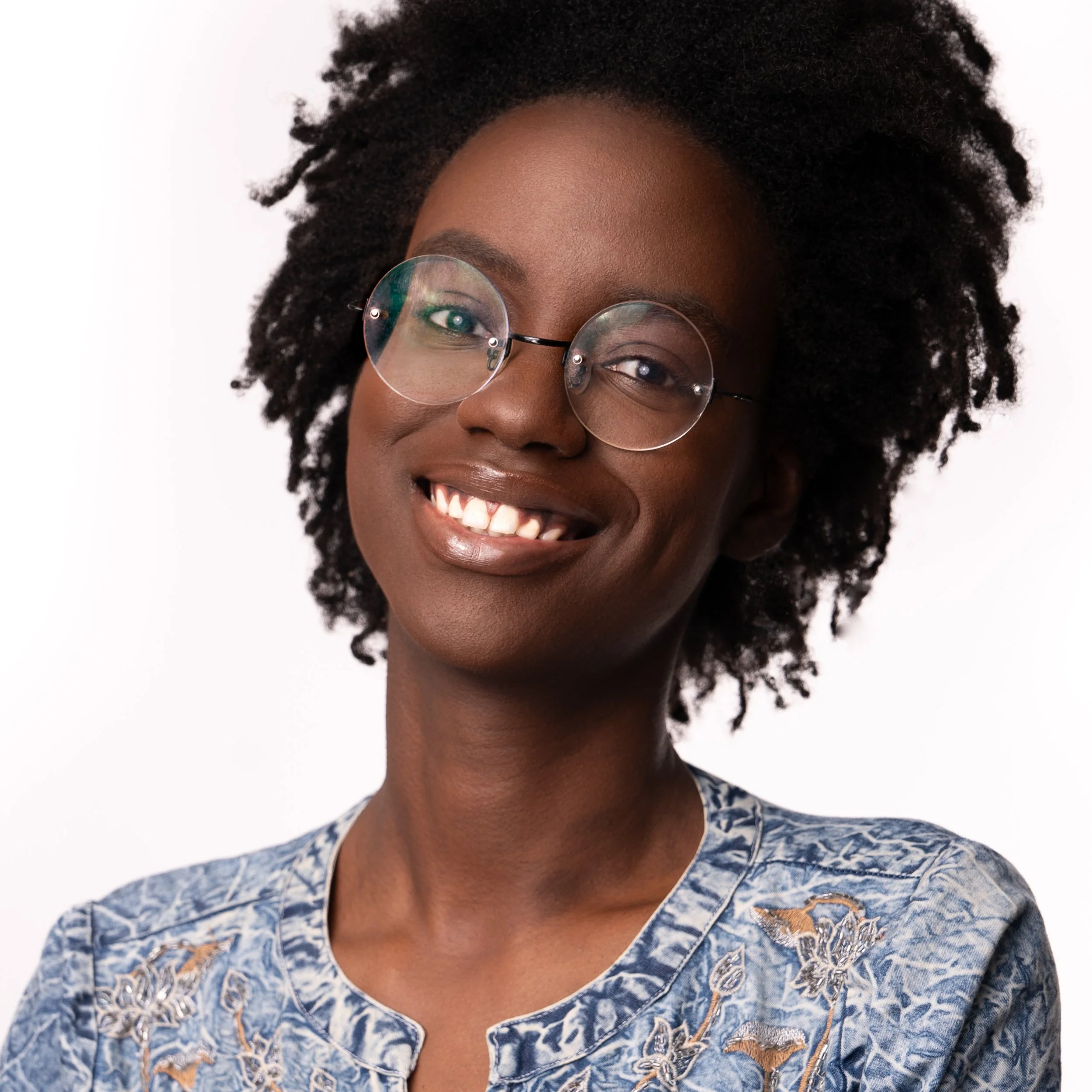A smiling Black woman with natural curly hair, wearing round glasses and a light blue embroidered top, against a white background.