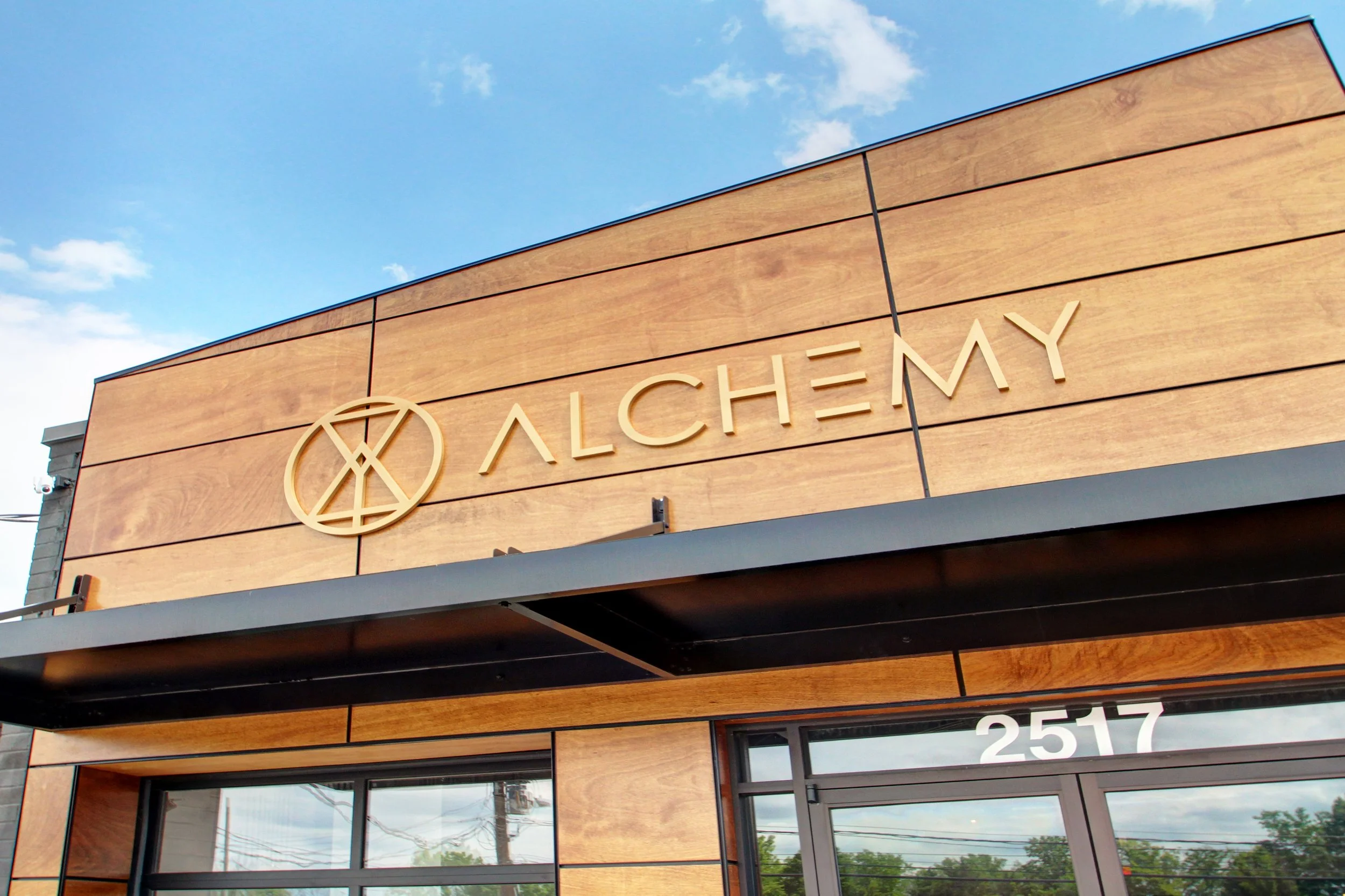 The exterior of a modern building with a wooden facade displaying the name 'Alchemy' and a logo, with the street number 2517 visible near the entrance and a partly cloudy sky in the background.