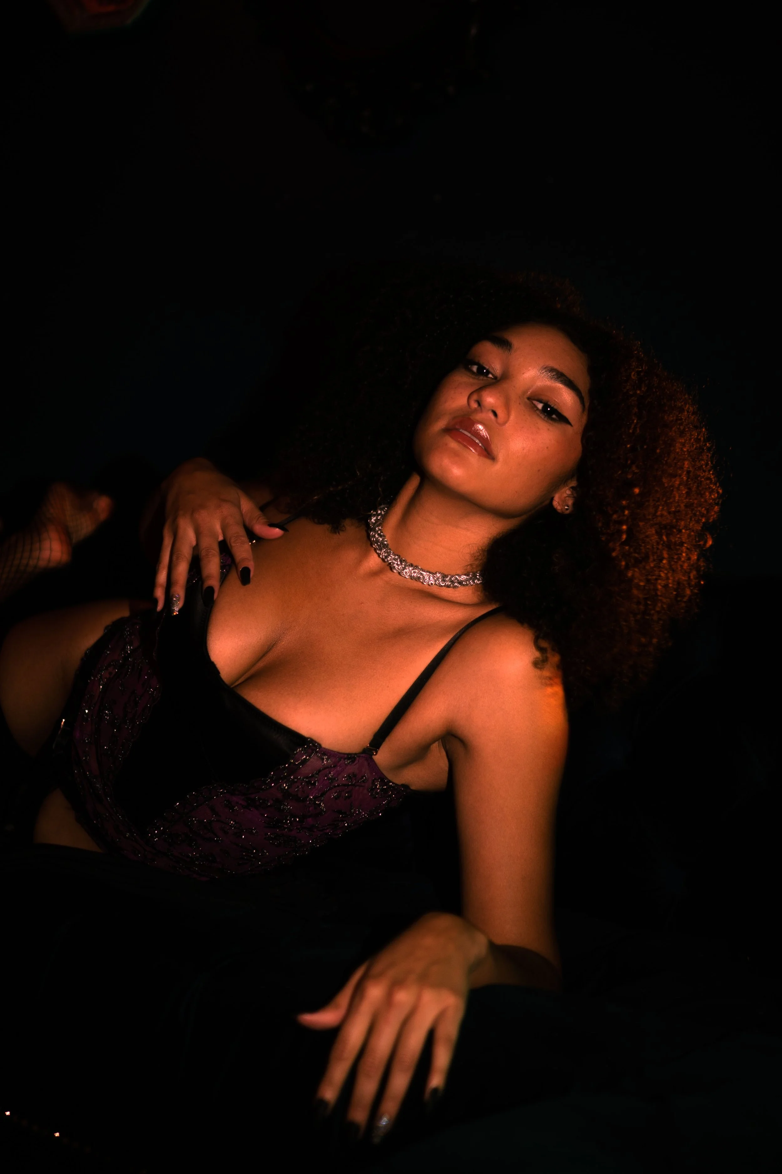 A woman with curly hair wearing a black and purple patterned top, silver necklace, and black nail polish, sitting in a dark environment.