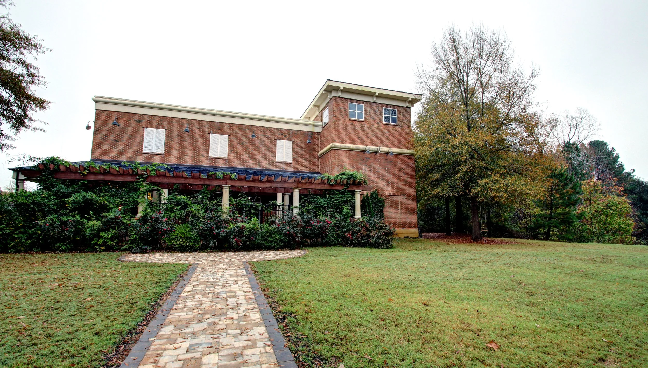 Brick house with a garden and a cobblestone pathway leading to the entrance, surrounded by trees with fall foliage.