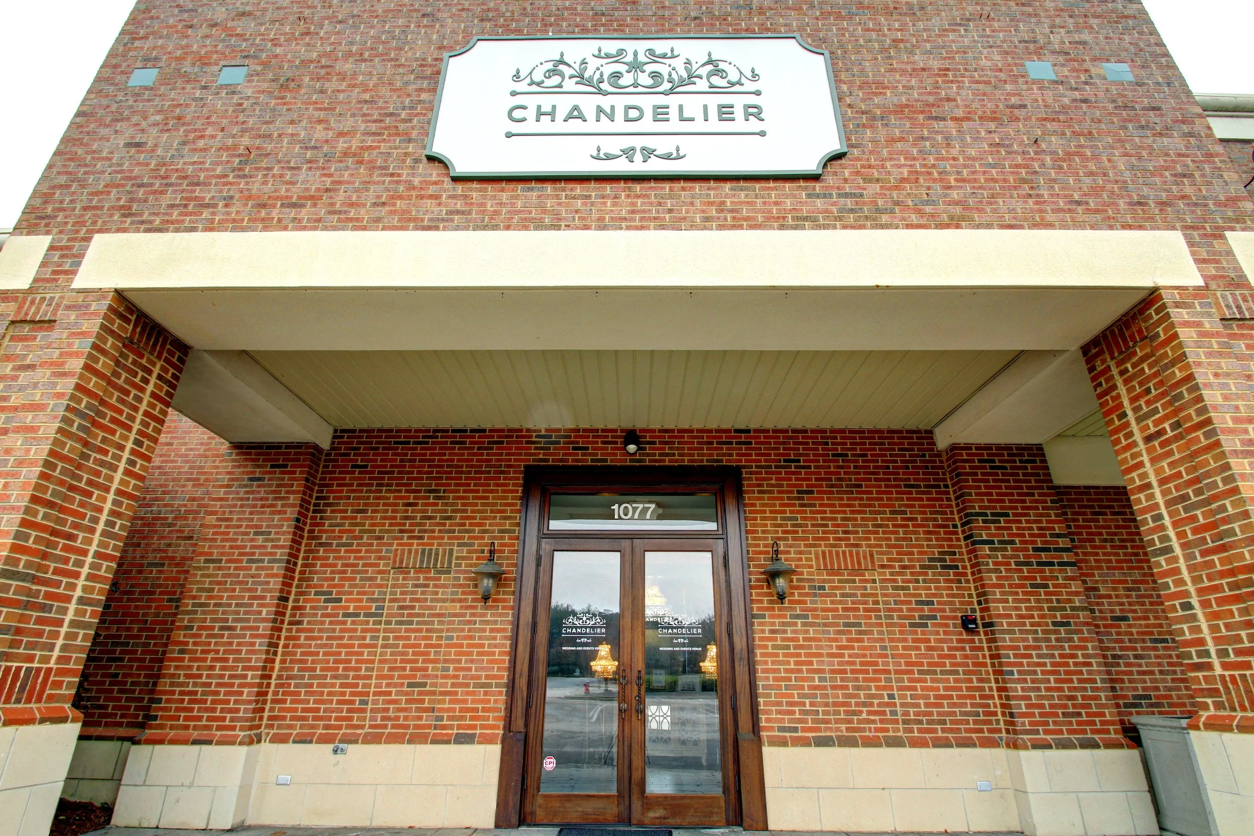 Brick building with a sign that reads 'Chandelier', a wooden glass door with the number 1077 above it, and two wall-mounted lamps flanking the door.