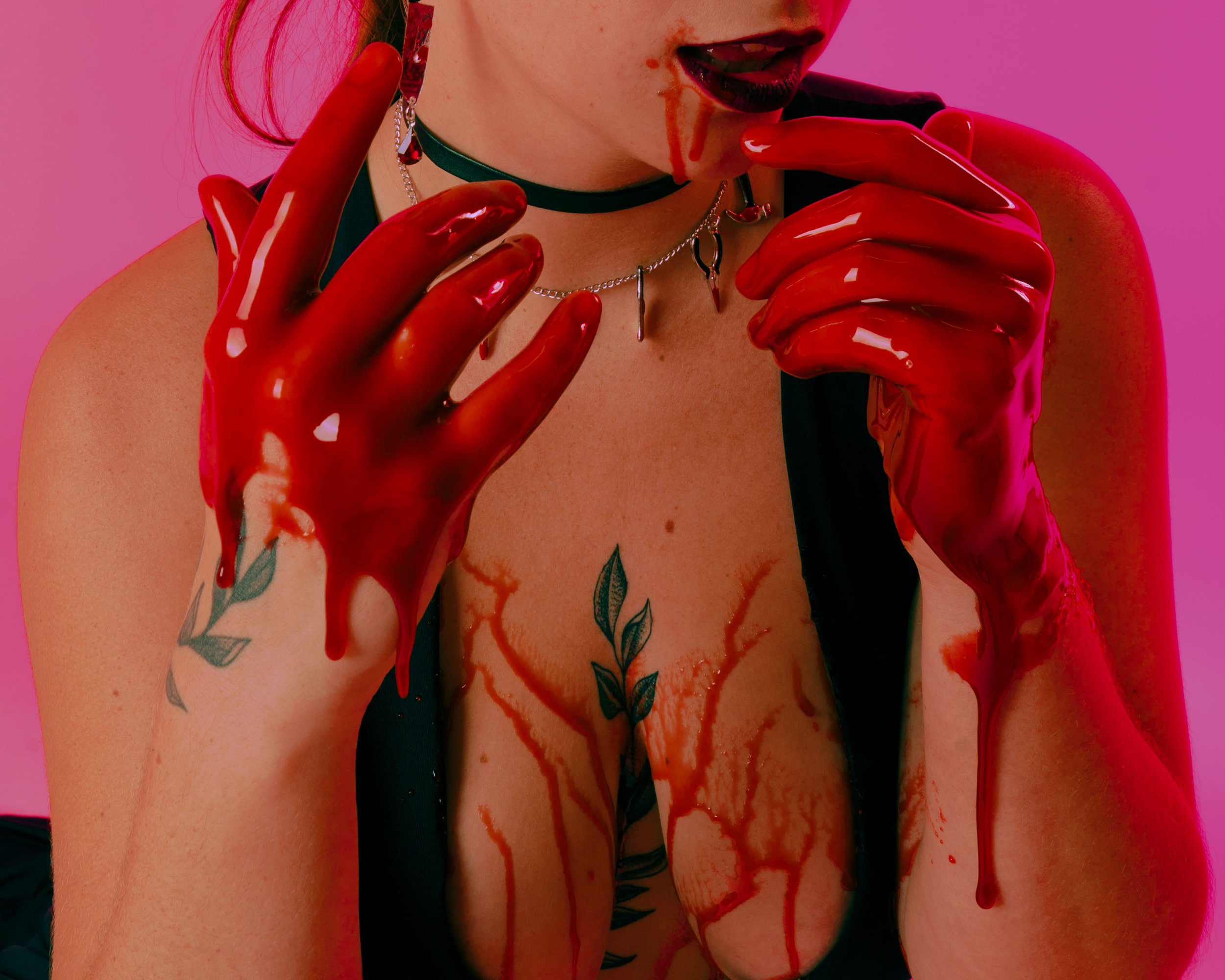 A woman with dark lipstick and tattoos on her chest and arm, wearing red latex gloves, is licking her fingers with blood around her mouth, set against a pink background.