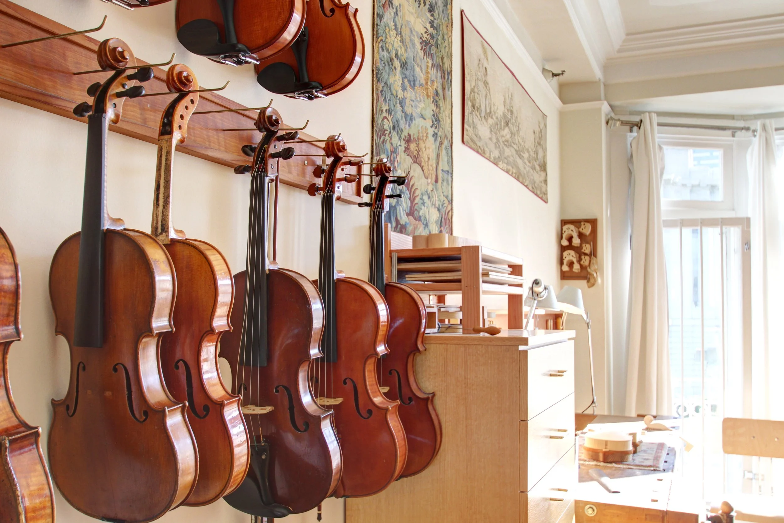 Multiple violins hanging on a wall in a room with wooden furniture, a tapestry, and a large window with white curtains.