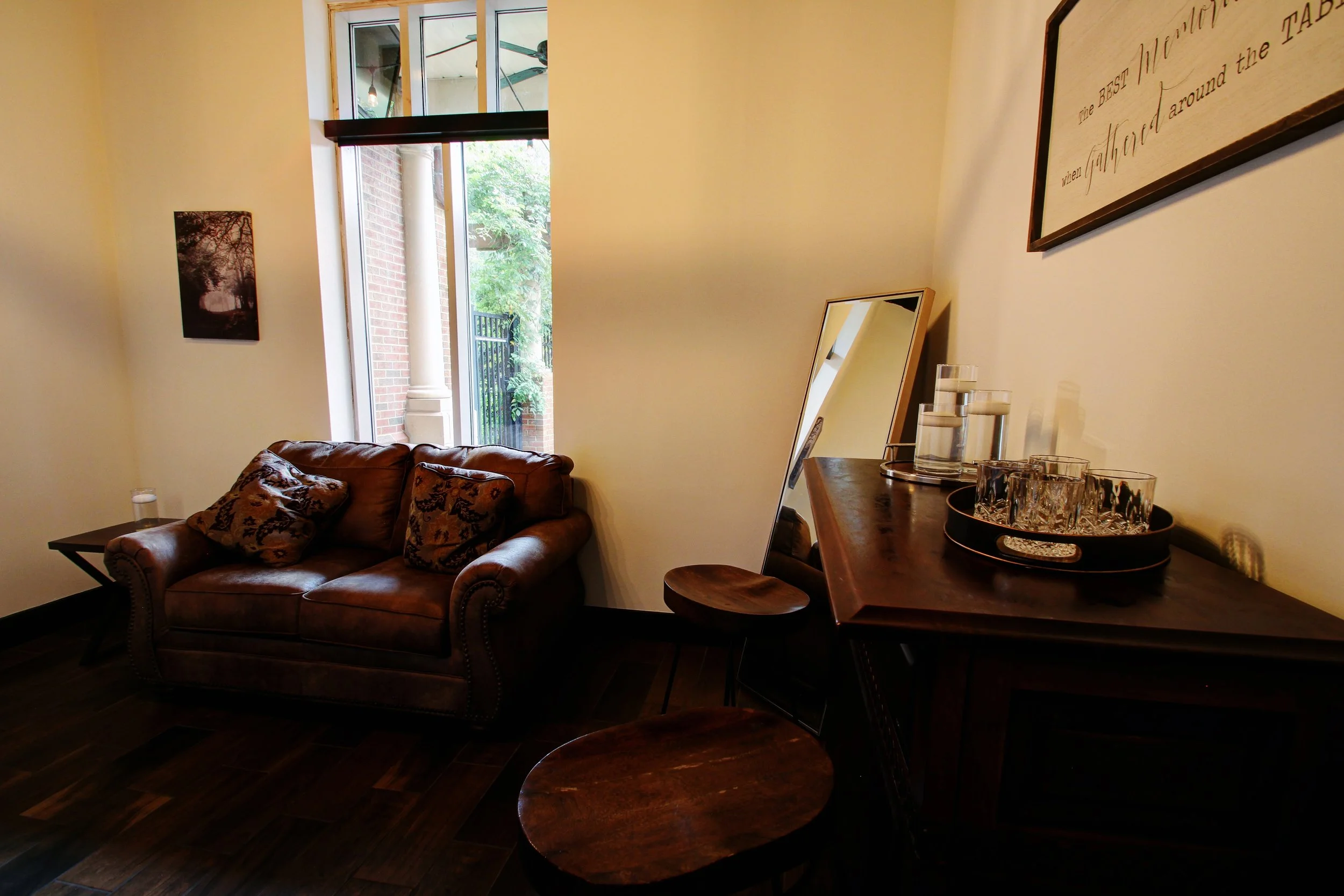 Interior view of a cozy living room with a leather sofa, decorative pillows, a small side table, a large mirror leaning against the wall, and a wooden sideboard with glassware. A window shows outdoor greenery.