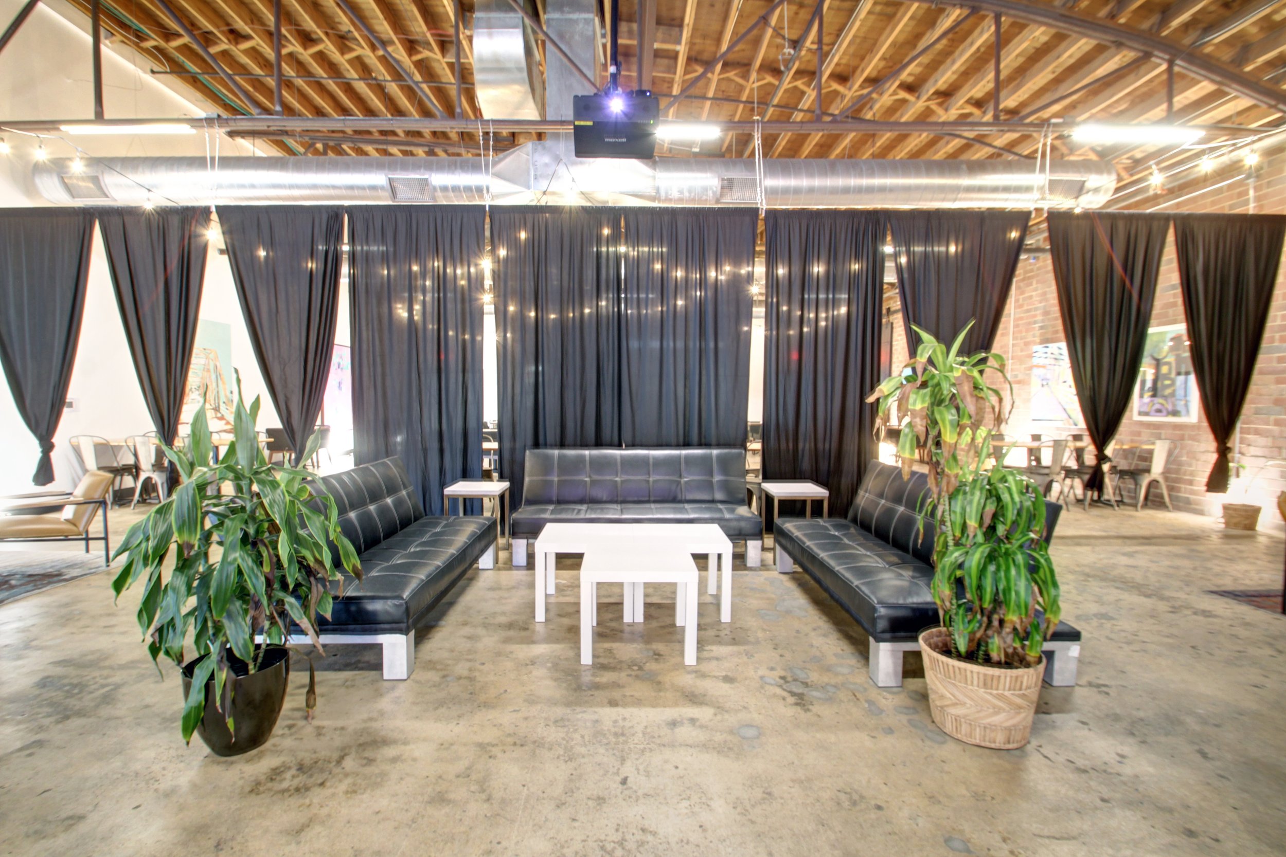 Indoor lounge area with black leather sofas, white tables, potted plants, black curtains, string lights, and a brick wall ceiling.