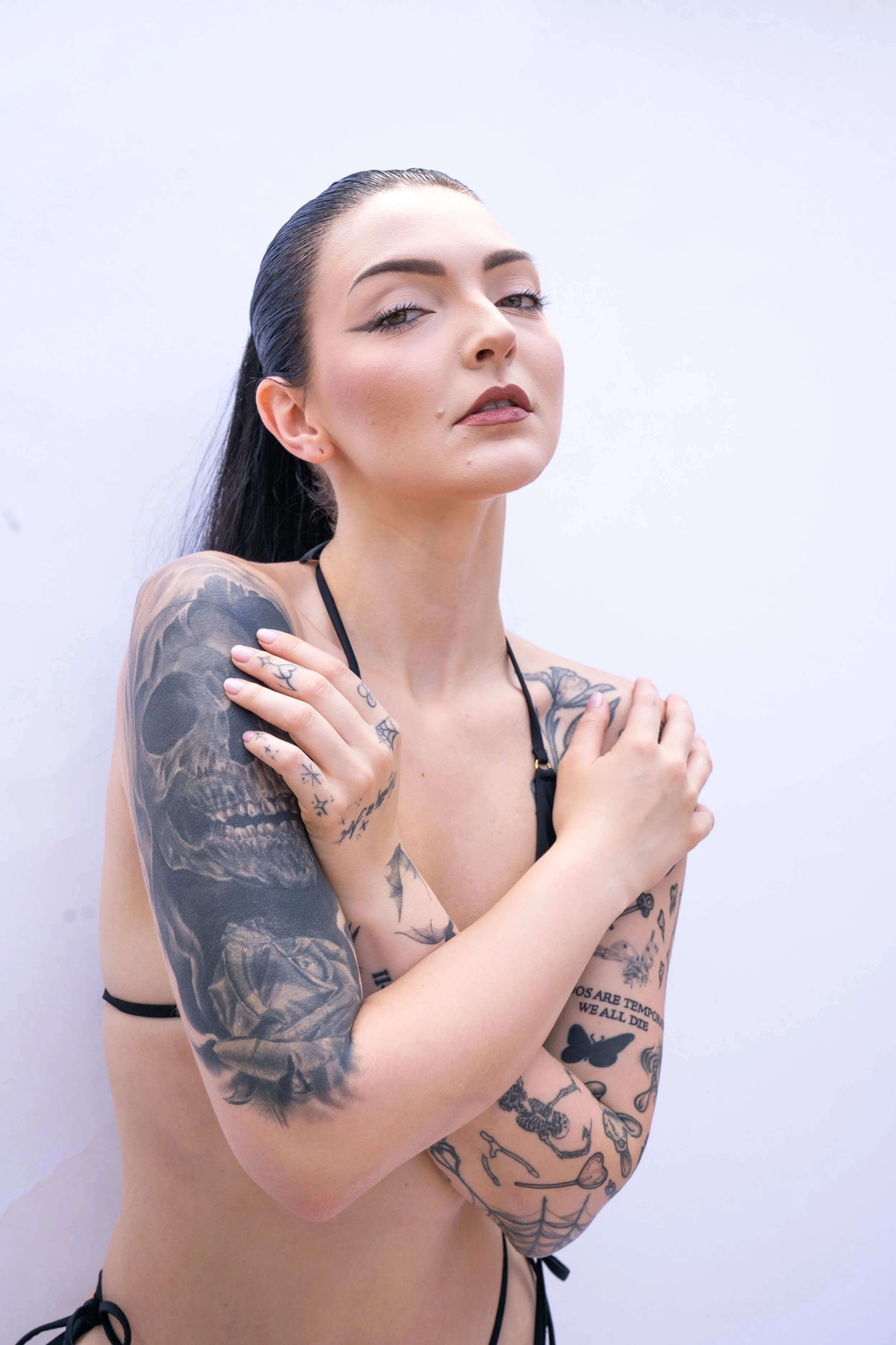 A woman with dark hair slicked back and tattoos on her arms and chest, crossing her arms over her shoulders and looking at the camera against a plain white background.