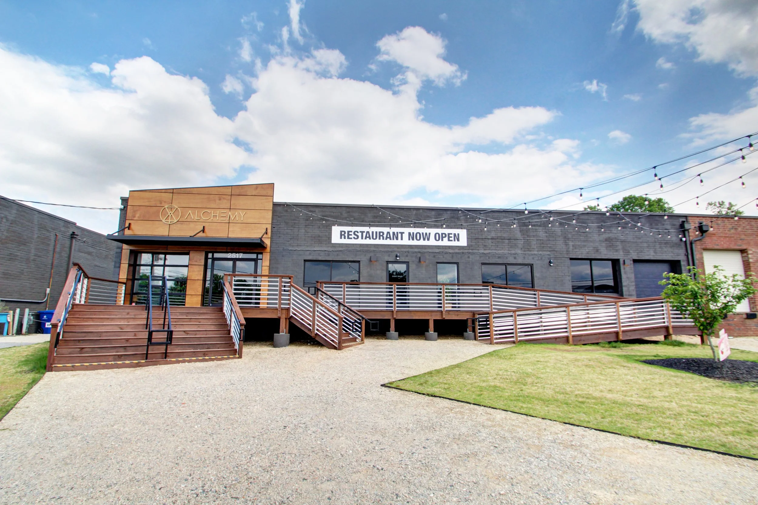 Exterior view of a modern restaurant building with a sign that reads 'Restaurant Now Open,' wooden stairs, a deck, and a small landscaped yard with grass and young trees.