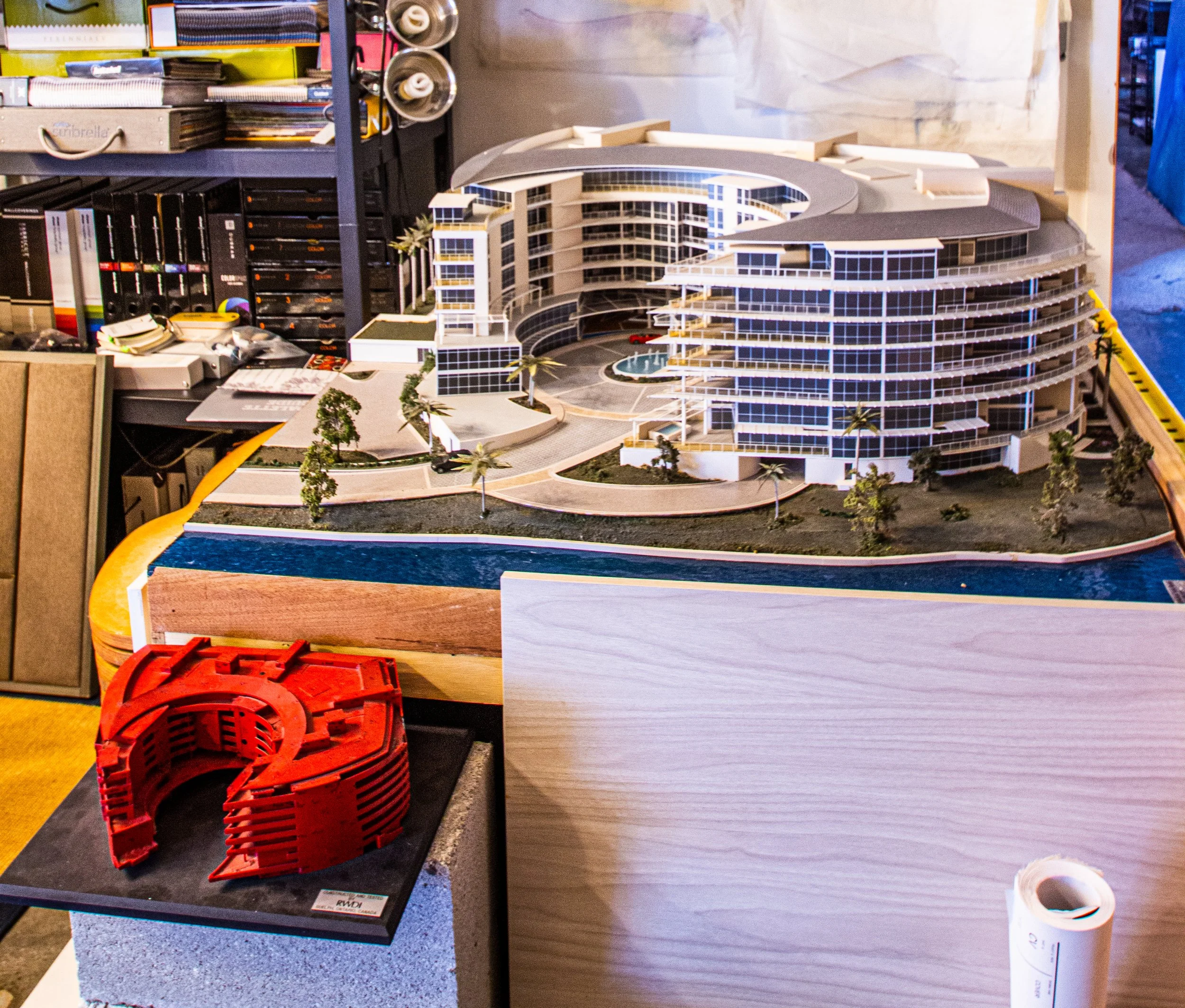 Architectural model of a modern multi-story building with curved design, surrounded by trees, and a water feature.