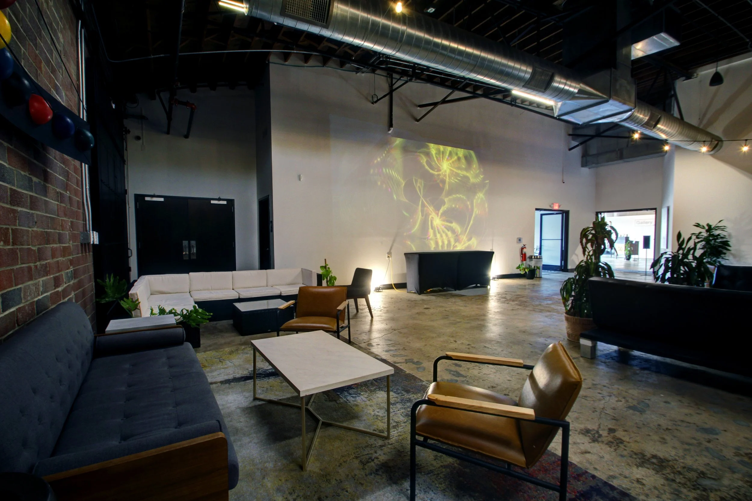 Modern lounge area with various couches, chairs, and plants, set in an industrial-style space with exposed ductwork, a video projection on the wall, and an open entrance to the outside.