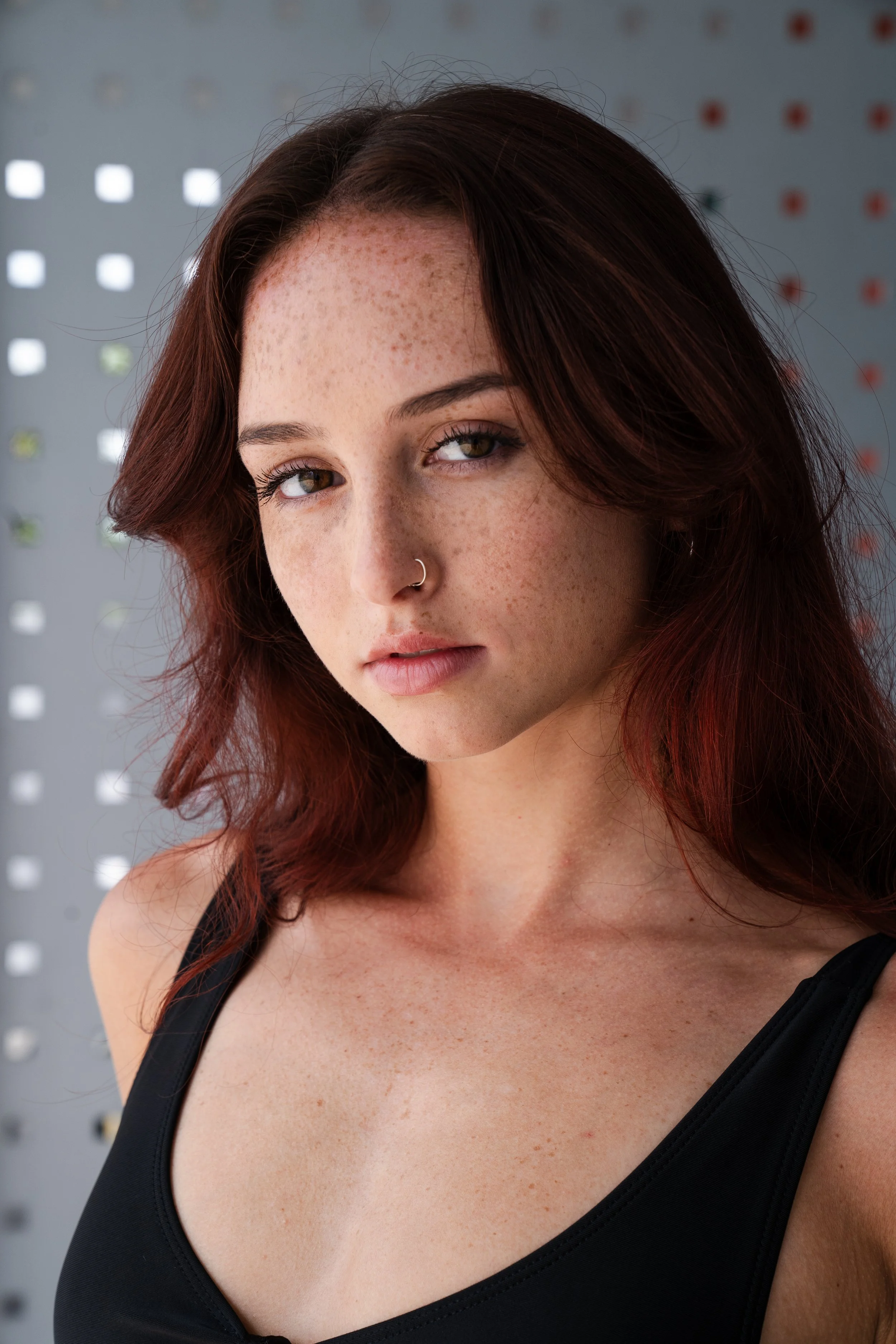 A young woman with reddish-brown hair, freckles, and a nose ring wearing a black tank top, standing in front of a perforated wall.