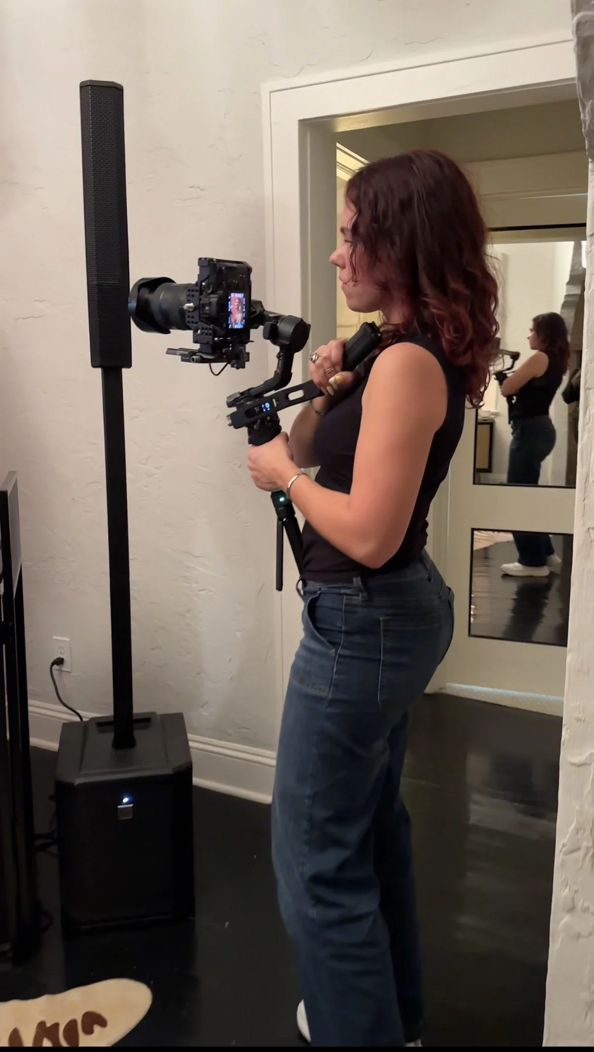 Woman with red hair filming with a professional camera mounted on a stabilizer at an indoor location, with a mirror in the background reflecting her.