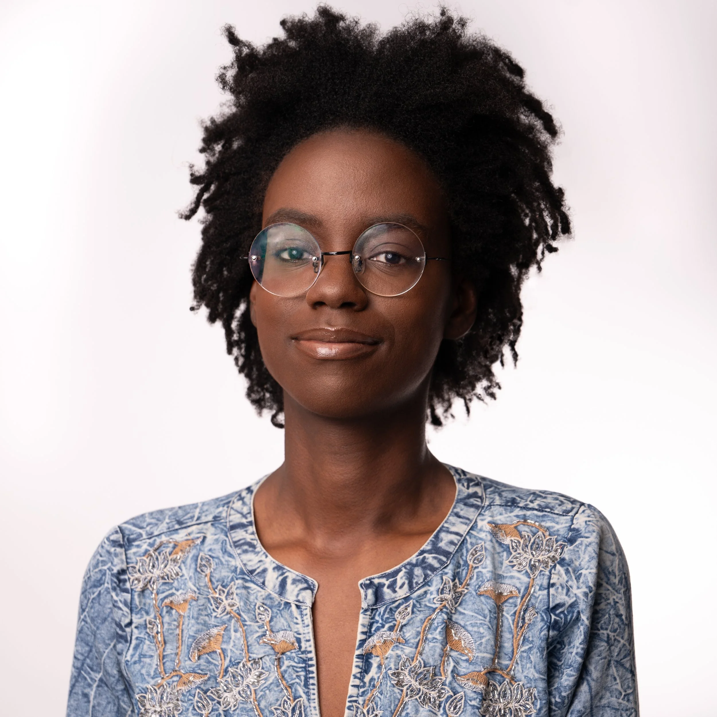 Portrait of a Black woman wearing round glasses and a blue embroidered top, smiling softly against a white background.