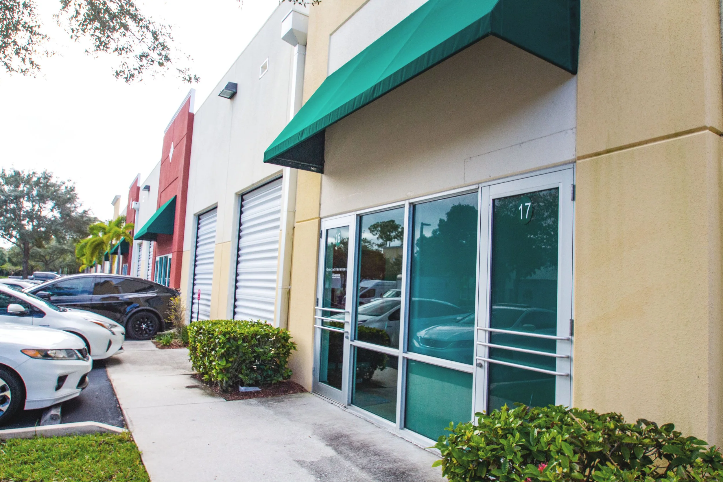 Image of a commercial building with green awnings, a glass door, and a parking lot with parked cars.