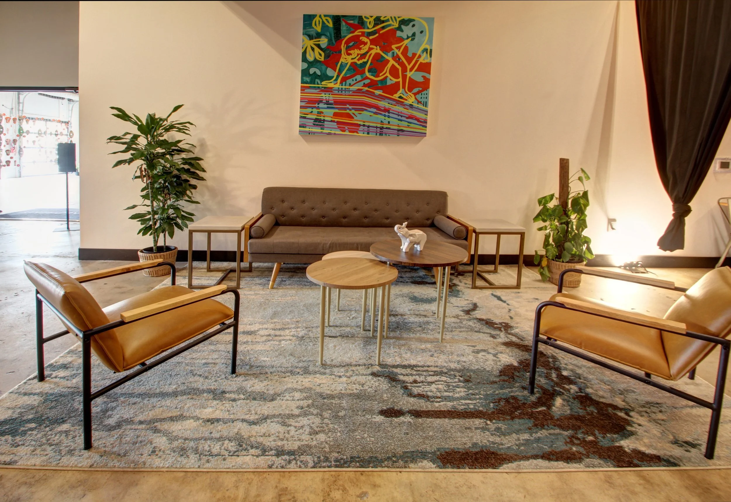 Modern living room with beige and leather furniture, including a brown sofa with tufted back, two leather armchairs, and small wooden side tables. There are two potted plants, colorful abstract wall art above the sofa, and a patterned area rug on the