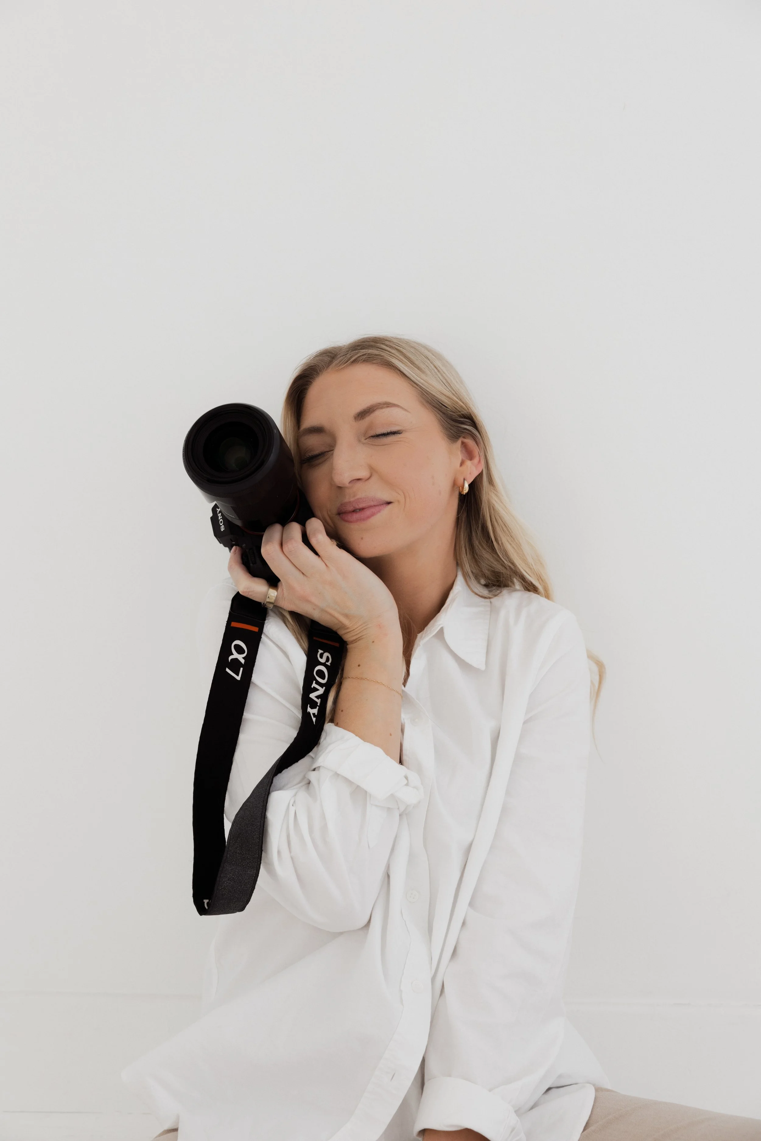 A woman with blonde hair and light skin, wearing a white shirt, is smiling gently with her eyes closed while holding a Sony camera with a large lens close to her face, against a plain white background.