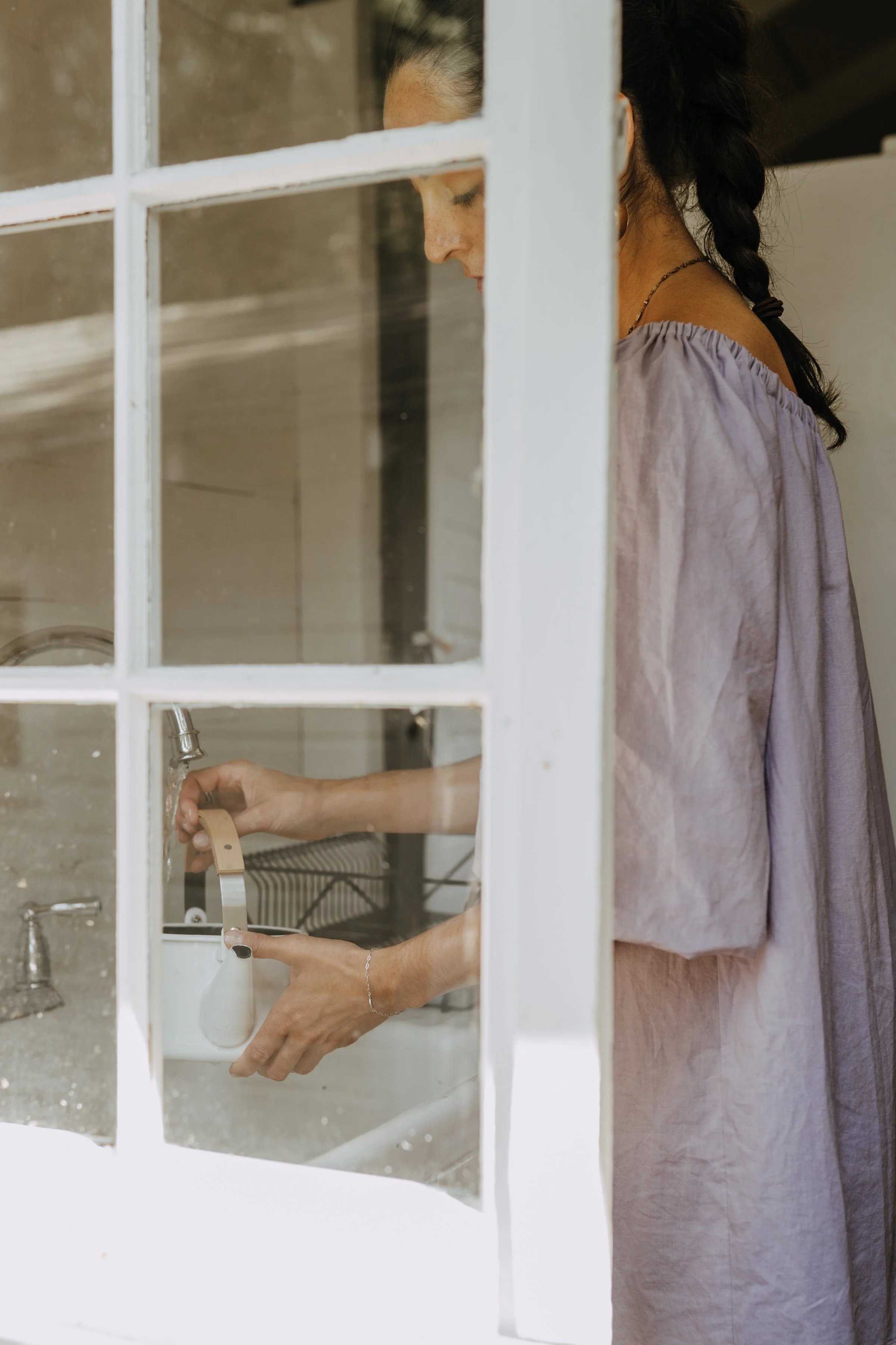Woman with dark hair tied in a braid, wearing a purple top, seen through a window, filling a soap pump with liquid soap in a kitchen.