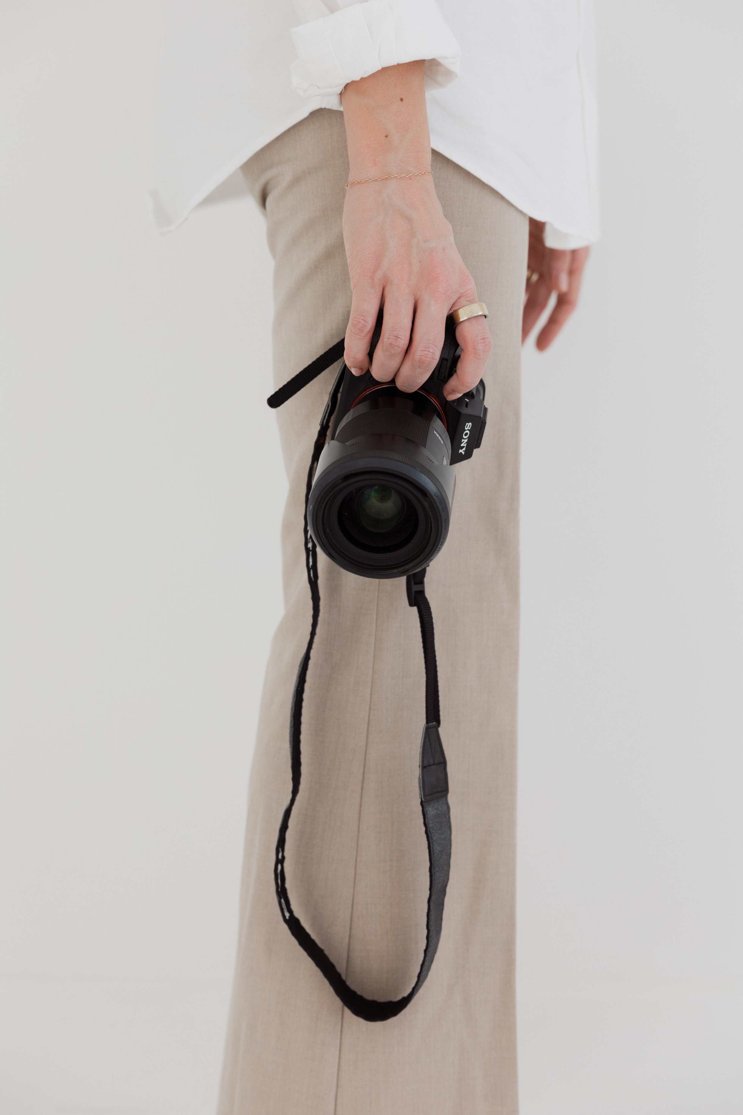 Person holding a professional camera, dressed in beige pants and a white shirt, with jewelry including a bracelet and a ring.