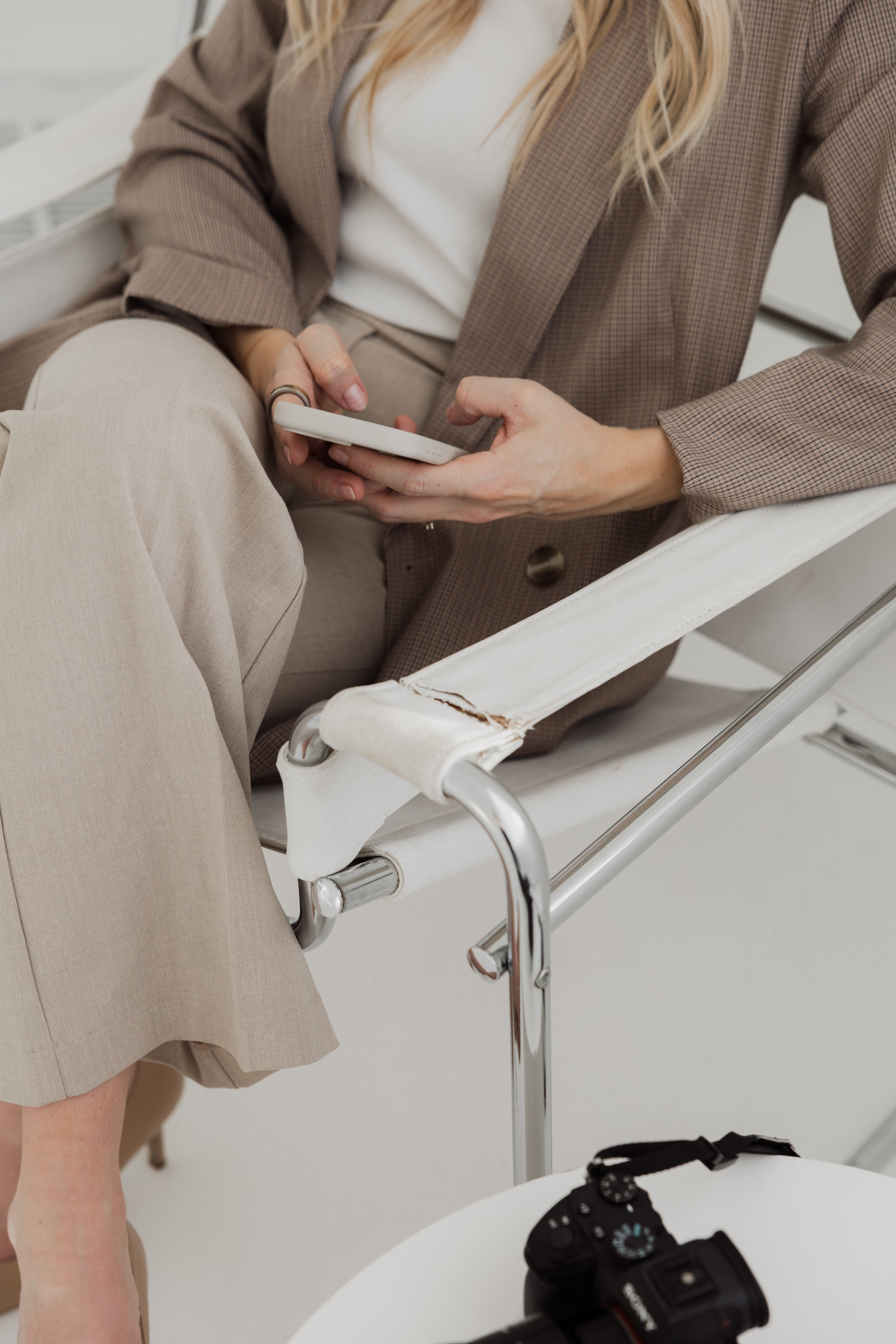 A woman sitting on a hospital bed or examination chair, dressed in beige pants and a brown checkered blazer, holds a smartphone in her hands and is looking at it. A black camera is placed on a white surface nearby.