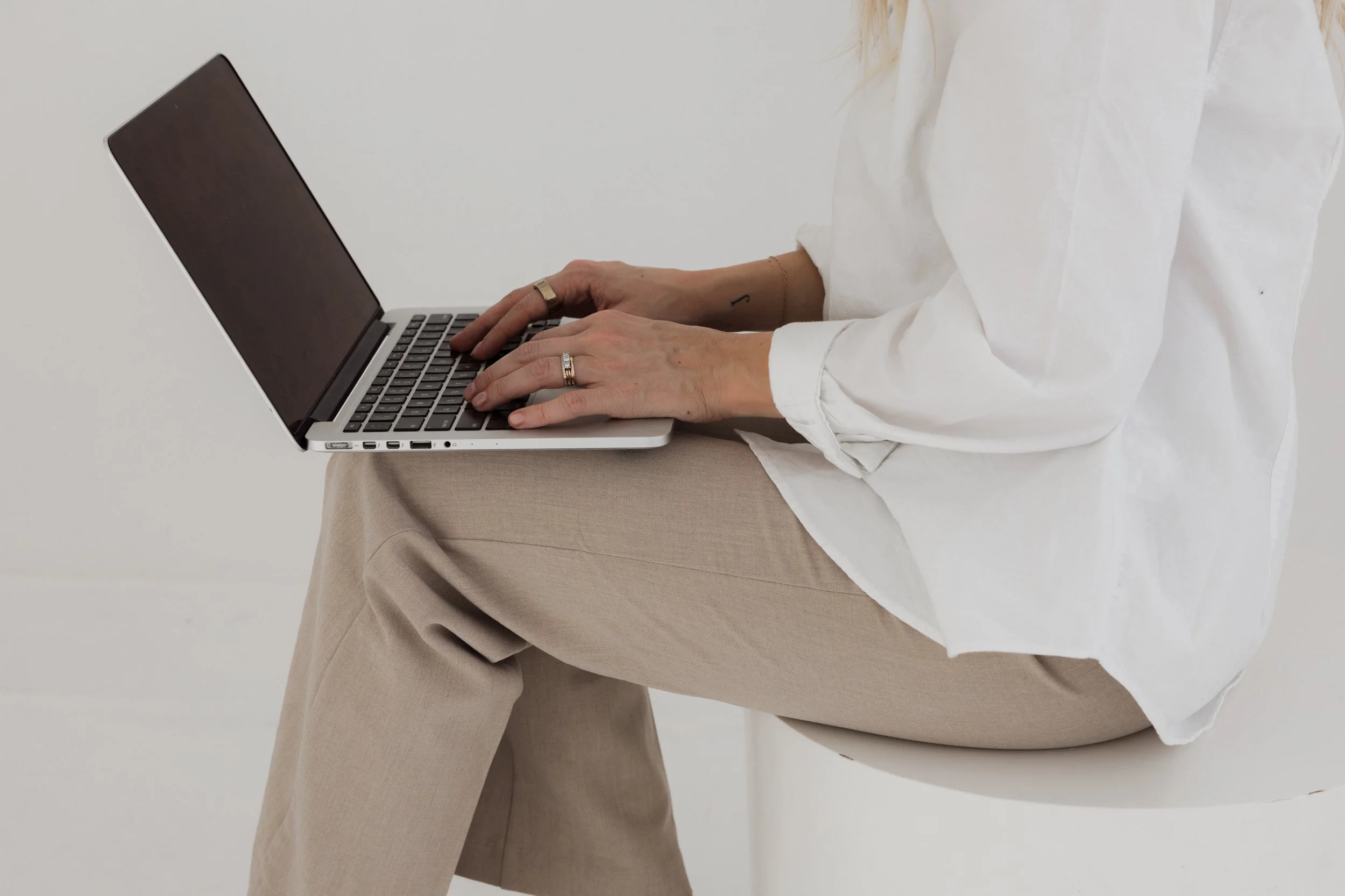 Person wearing a white shirt and beige pants working on a laptop, sitting on a white chair, with hands on the keyboard.