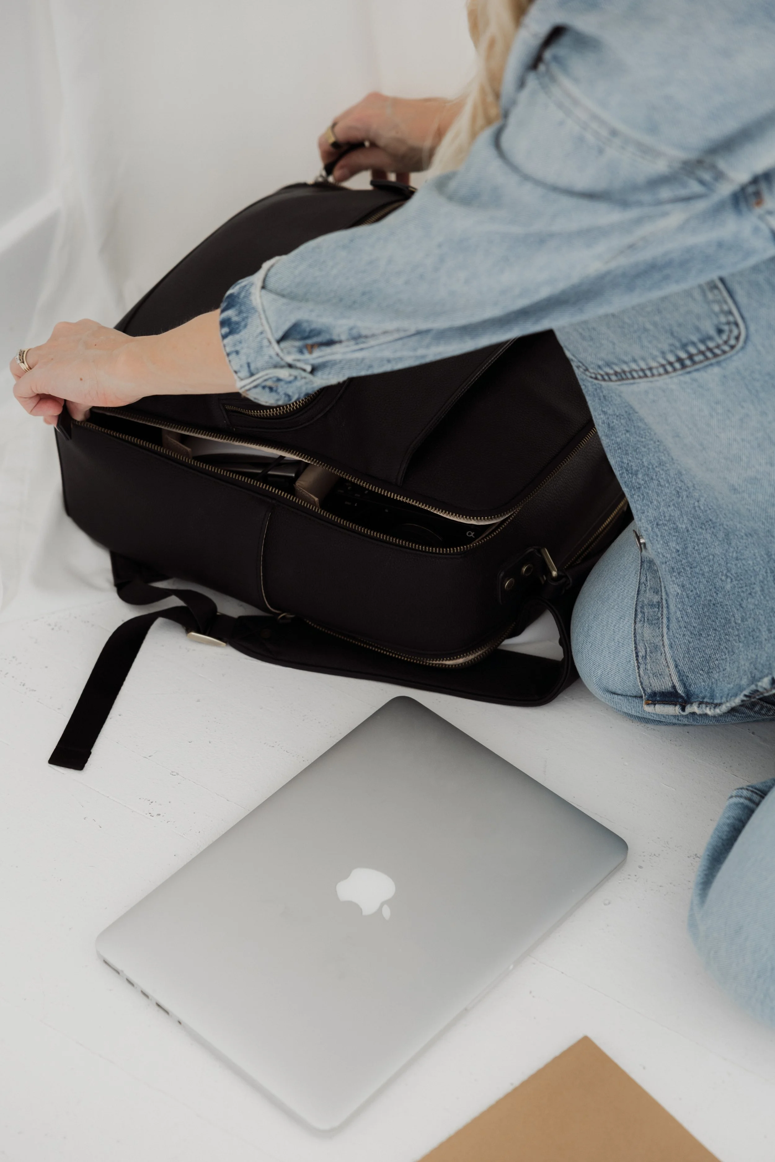 Person wearing a denim jacket packing a black bag, with a closed silver MacBook laptop on a white surface nearby.