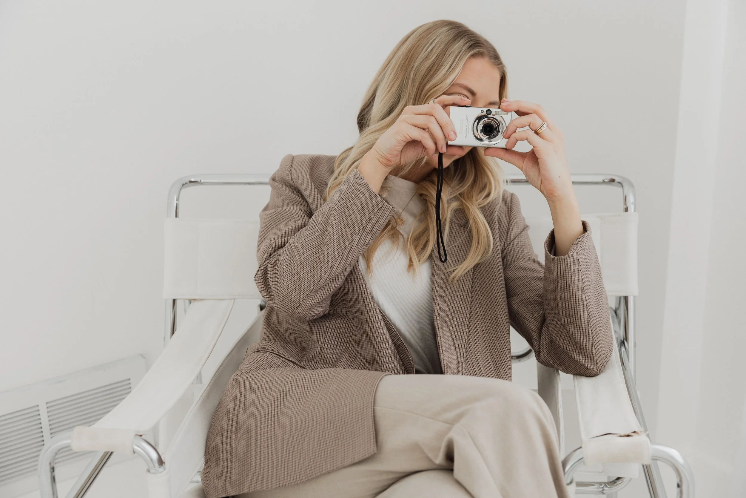 A woman with blonde hair wearing a beige blazer and light-colored pants taking a photo with a compact camera while sitting on a white chair in a minimalistic room