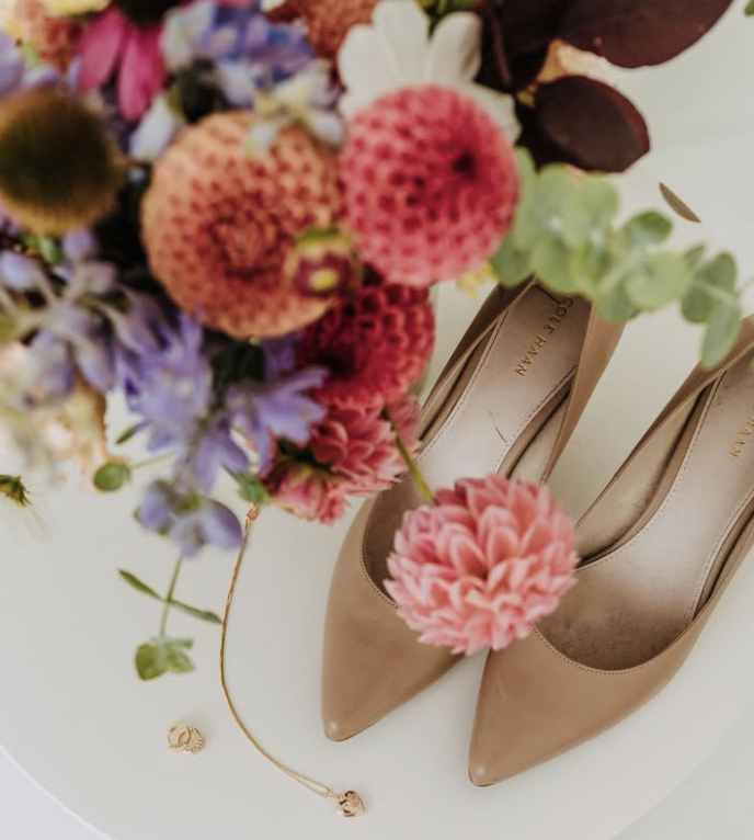 Beige high-heeled shoes next to a colorful bouquet of flowers on a white surface.