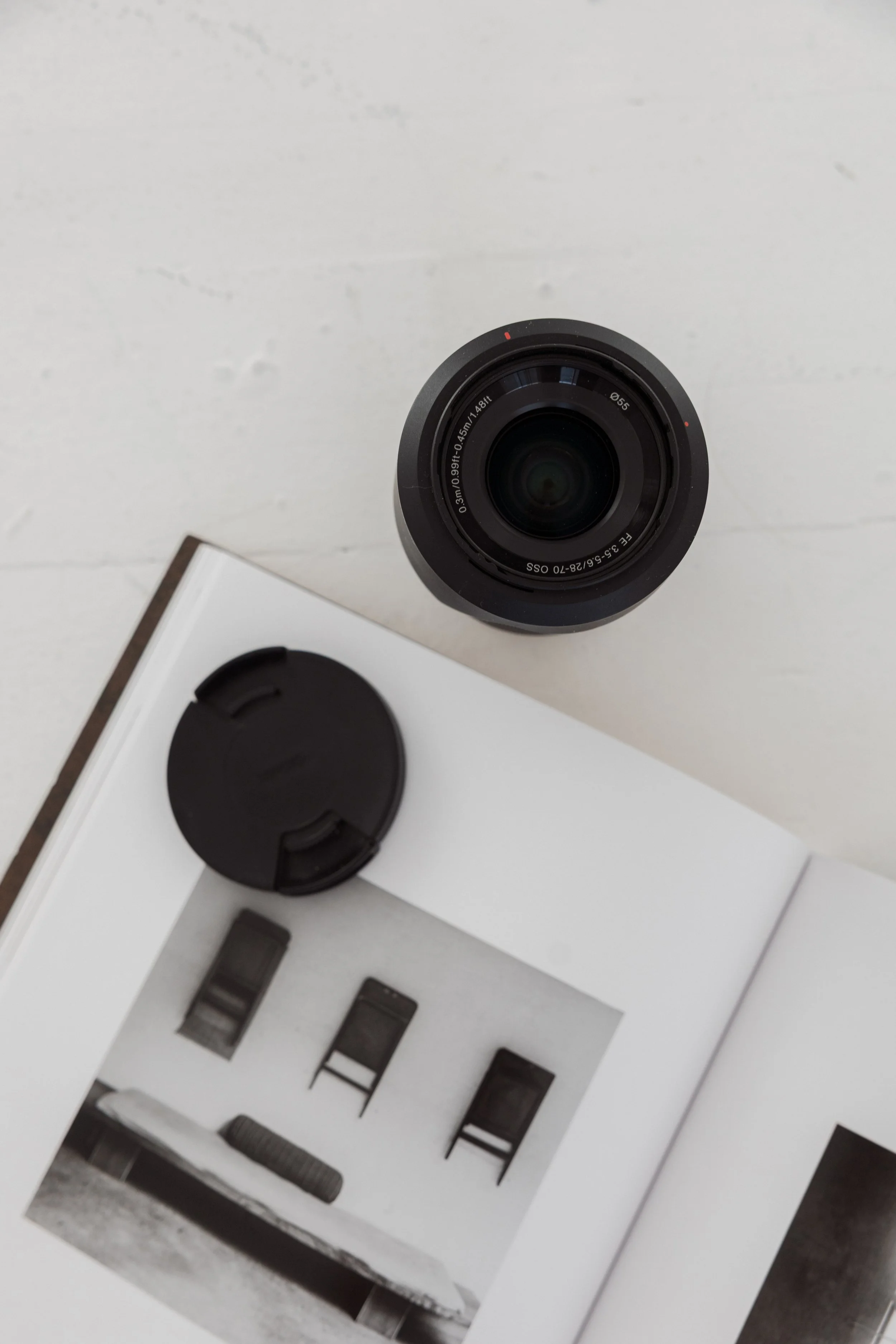 Top-down view of a camera lens, open book with black and white photos, and a camera lens cap on a white surface.