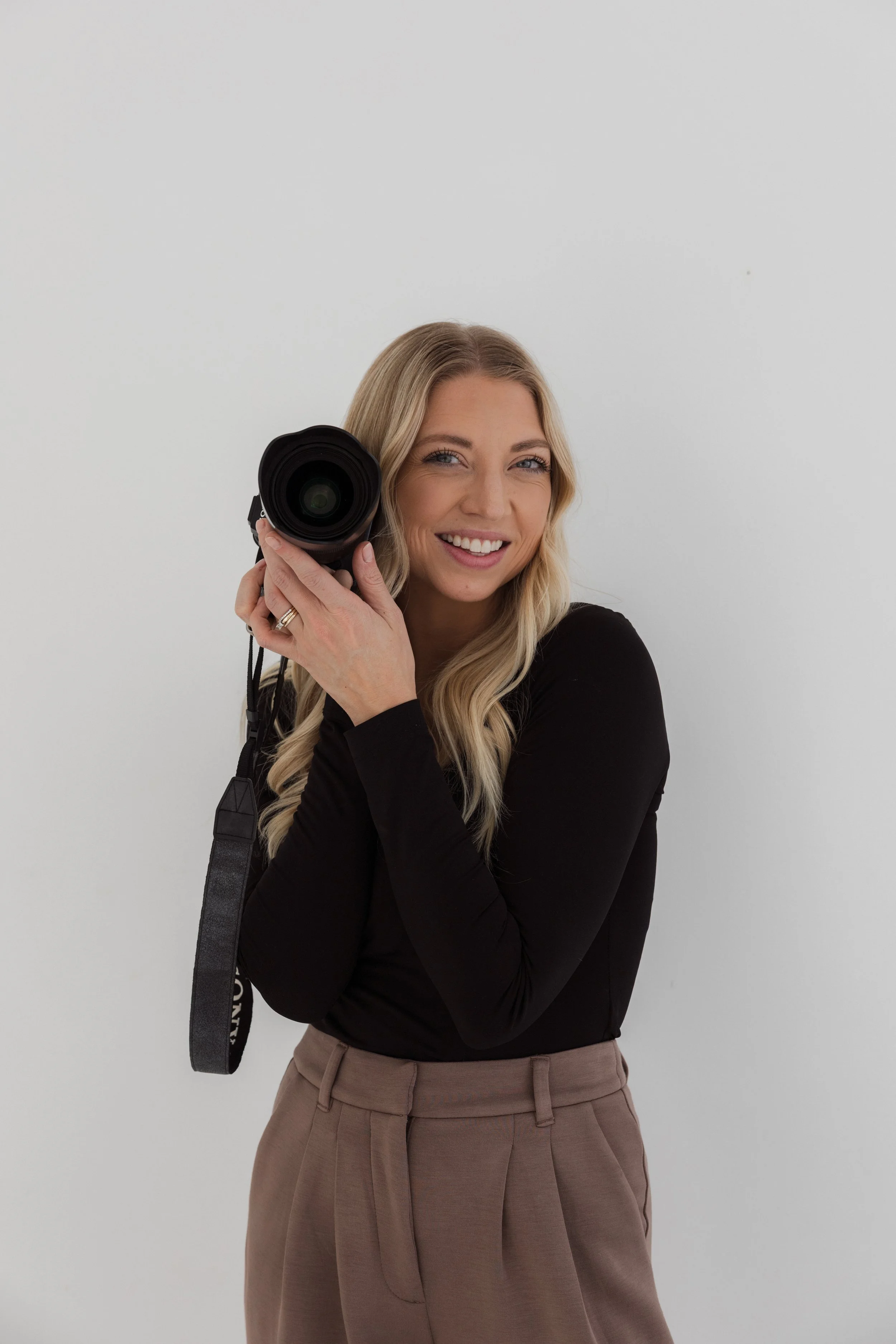 A woman with long blonde hair, wearing a black long-sleeve shirt and beige pants, smiling and holding a camera up to her face, standing against a plain white background.