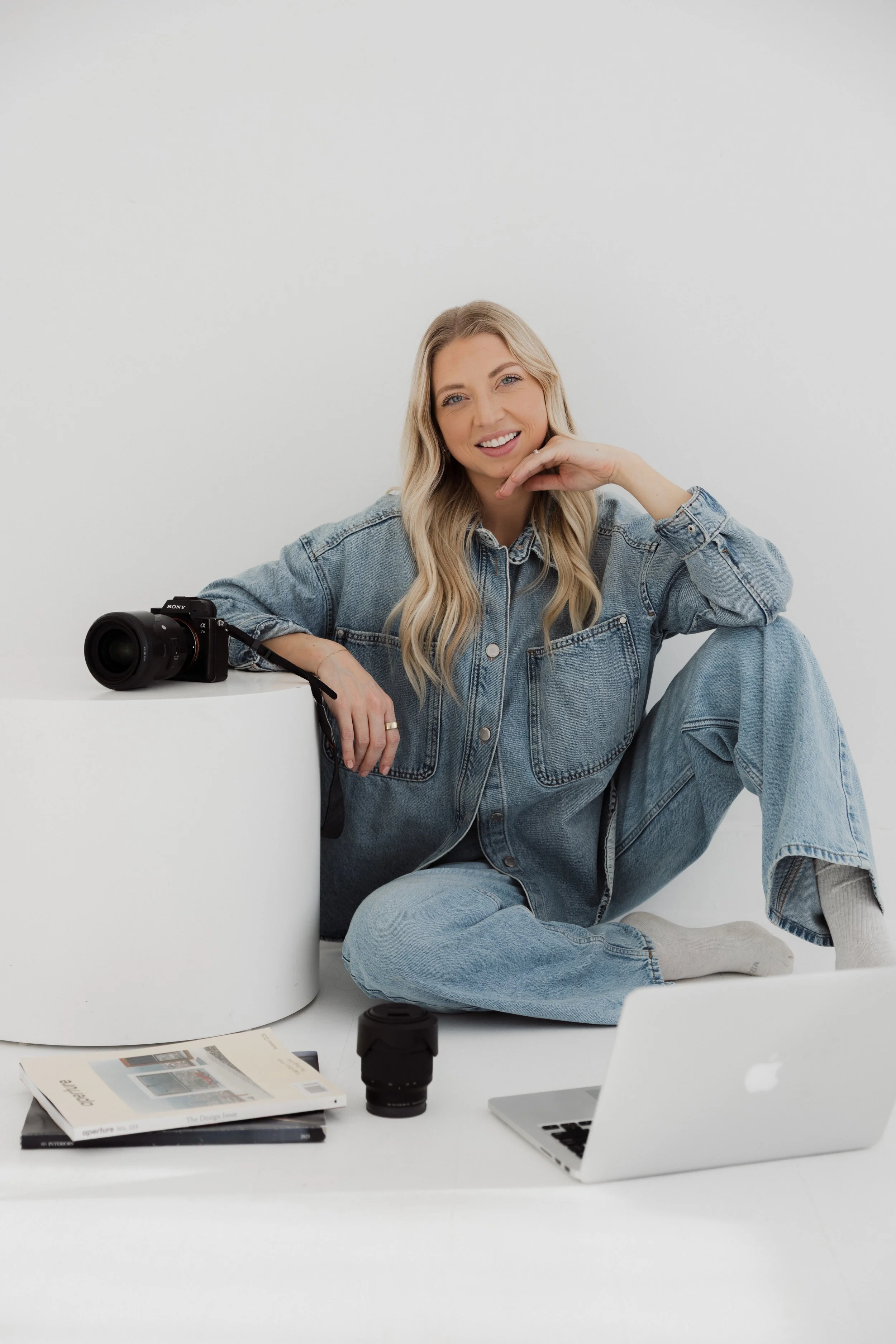 A woman with long blonde hair, wearing a denim jacket and jeans, sitting on the floor against a white wall, smiling and resting her chin on her hand. There is a camera on a white cylindrical table, a camera lens, some magazines, a camera lens cap, and a silver laptop in front of her.
