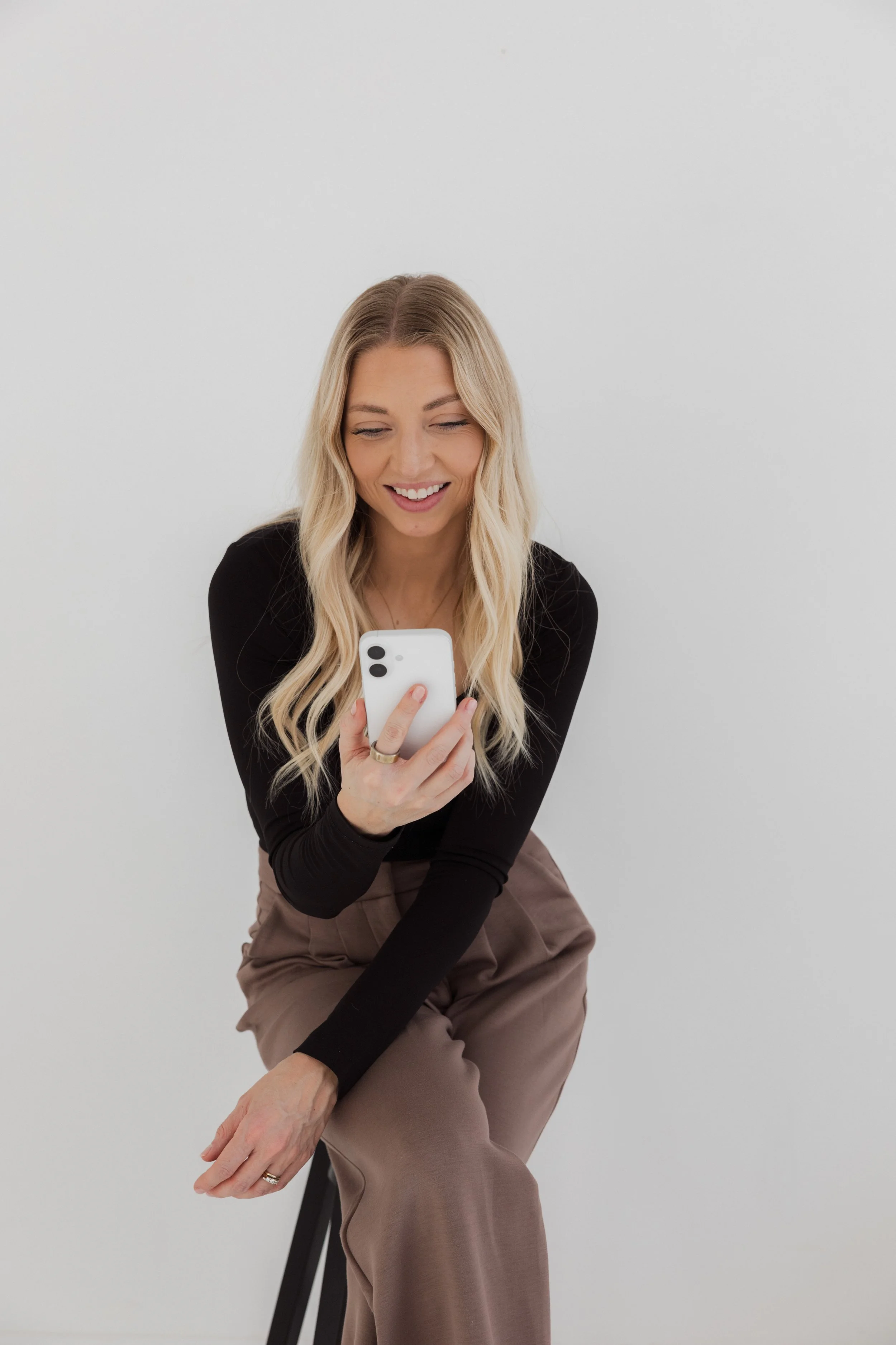 A woman with long blonde hair smiling while looking at her phone, sitting on a stool against a plain white background.