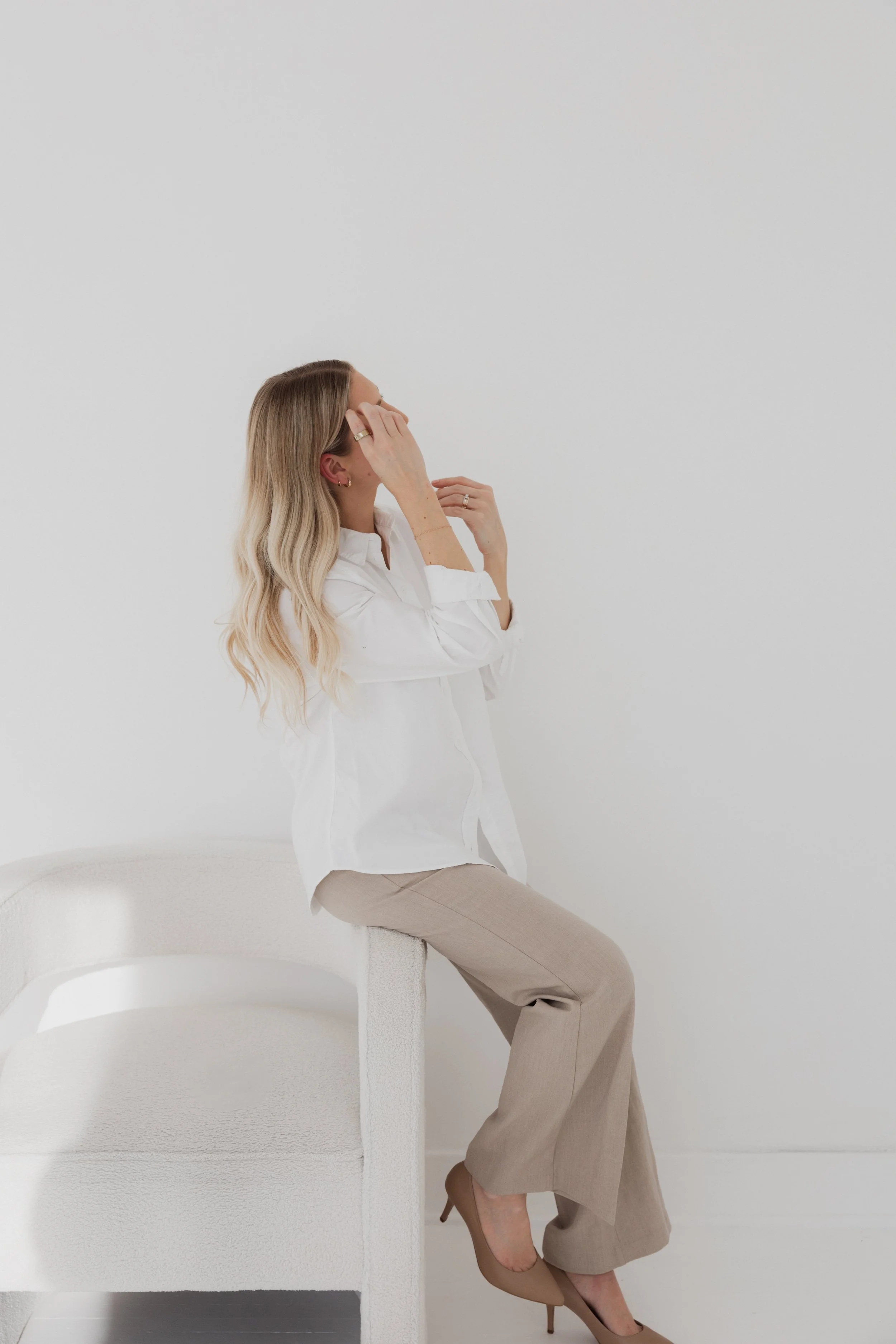A woman with long blonde hair wearing a white shirt and beige pants sitting on the armrest of a white textured sofa in a minimalist, white room, adjusting her earring.