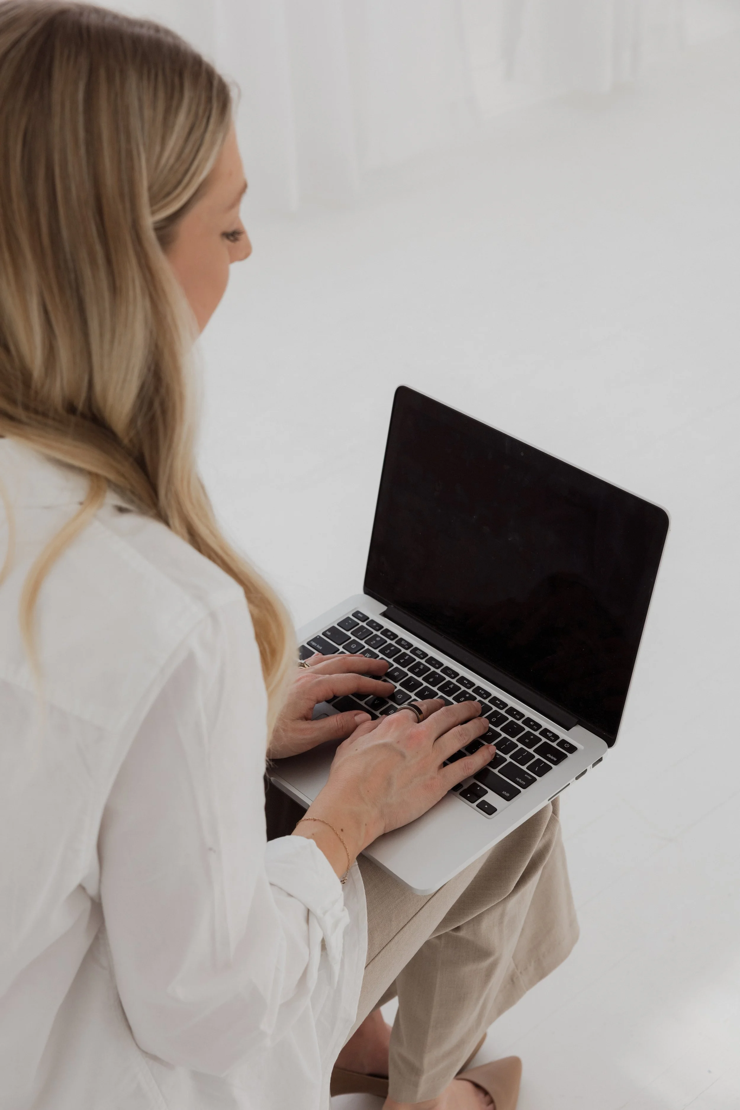 A woman with long blonde hair wearing a white blouse, sitting on a chair and typing on a silver laptop with a black screen.