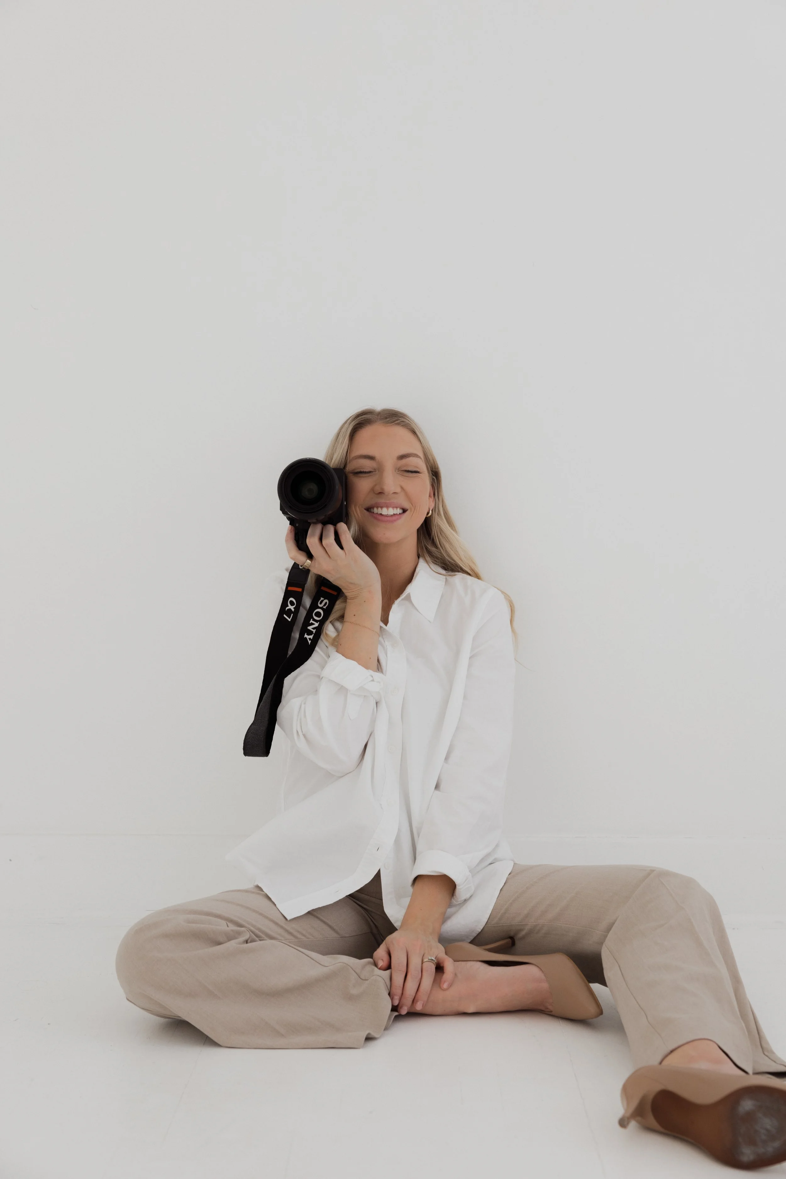 A woman sitting on the floor, smiling and holding a professional camera, wearing a white shirt, beige pants, and beige shoes, against a plain white background.