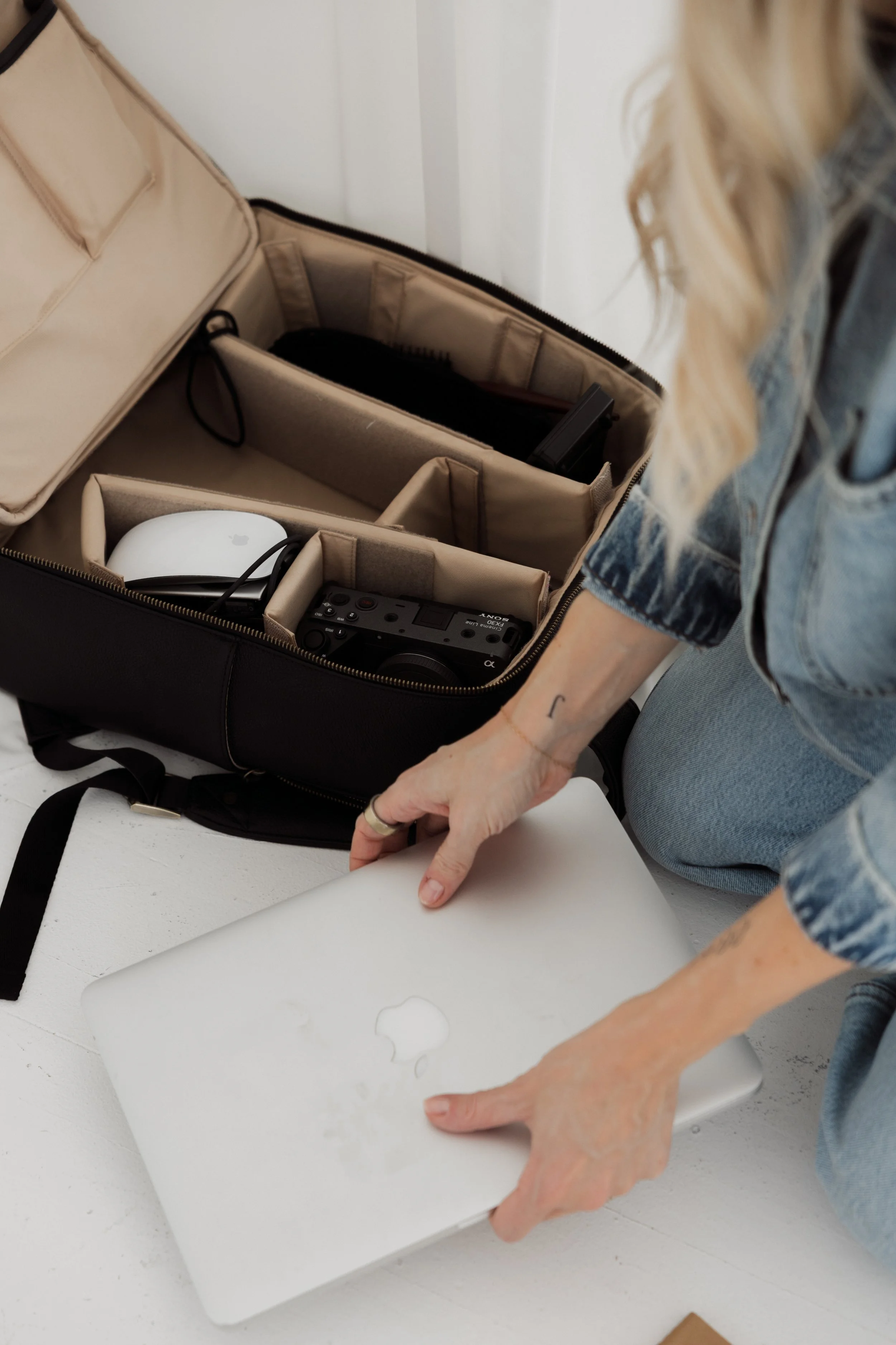 A woman placing a silver Apple MacBook laptop on the floor near an organized padded storage bag filled with electronic devices including a camera, a mouse, and other accessories.