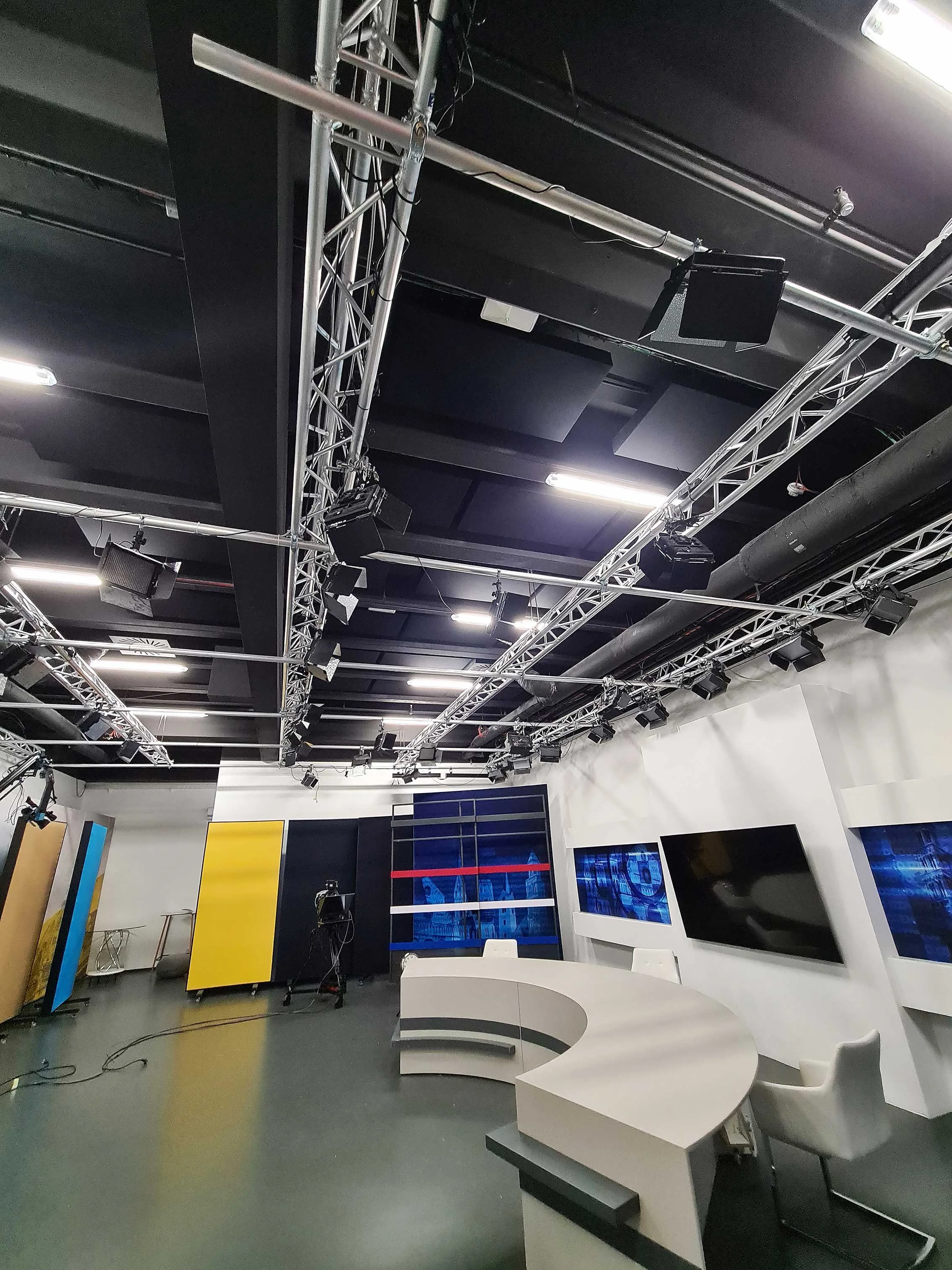 Television news studio with curved desk, white chairs, large monitors, and industrial ceiling with lighting and camera equipment.