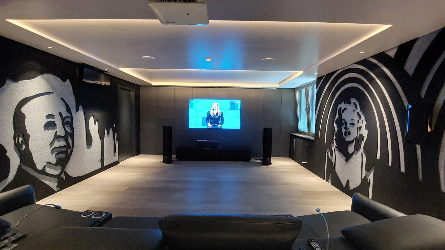 A modern living room with a dark wall featuring large black and white mural portraits of a man and a woman. There is a flat screen TV on the back wall displaying a woman, with speakers on either side. The ceiling has recessed lighting and a projector