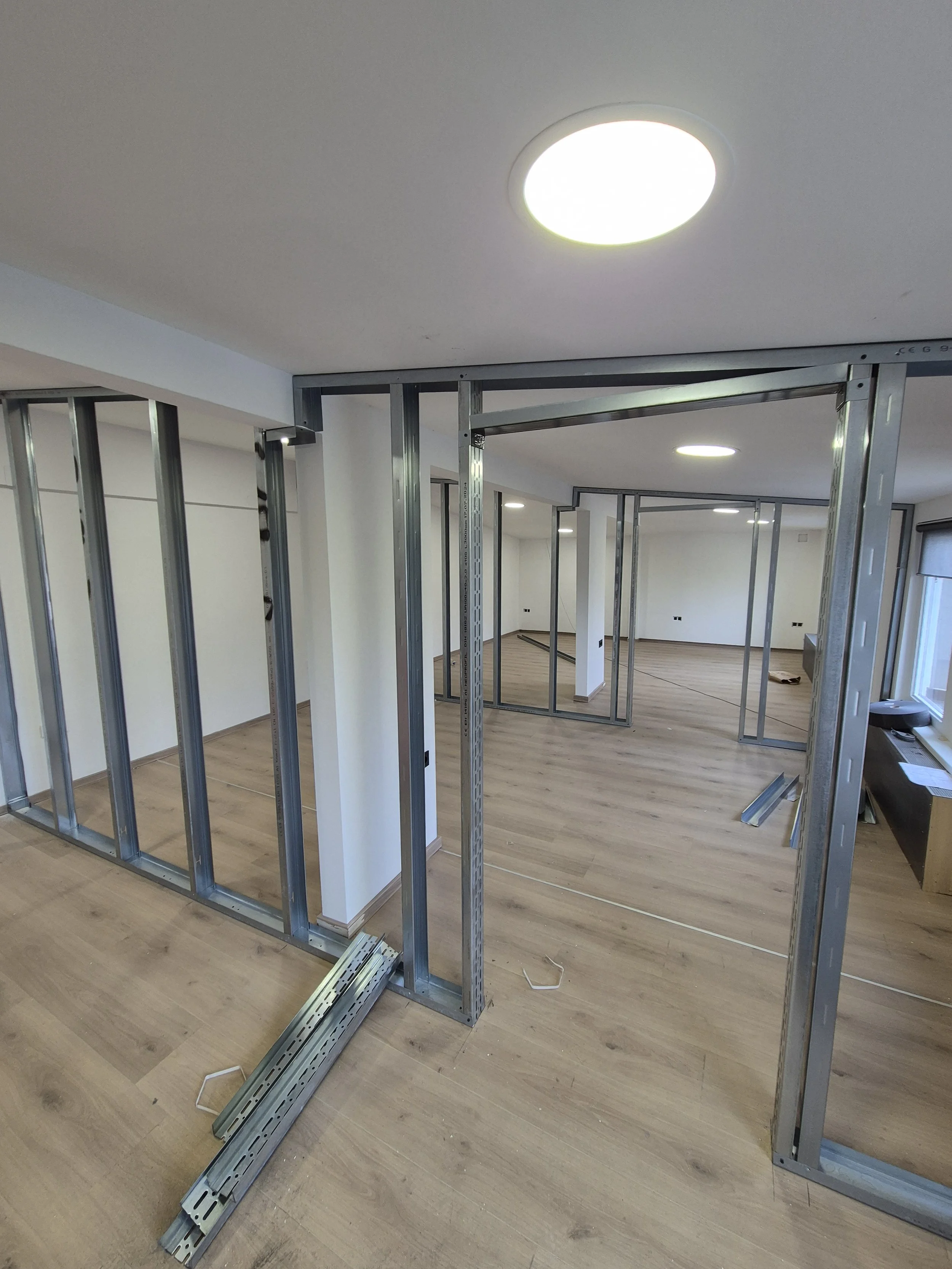 Interior of a room under construction with metal framing for walls, hardwood flooring, and ceiling lights.