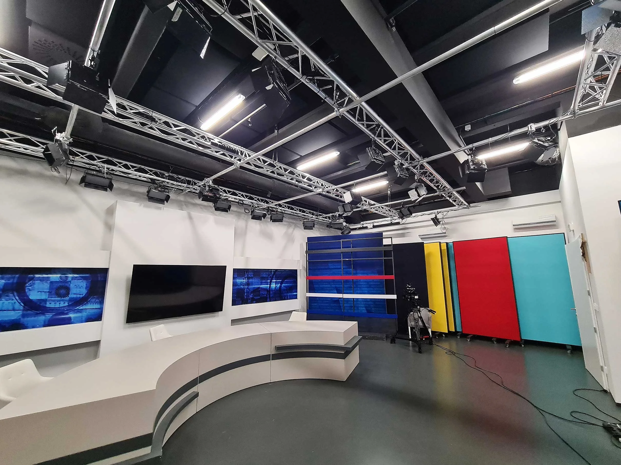 TV news studio with a curved desk, large screens on the wall, and colorful storage cabinets.