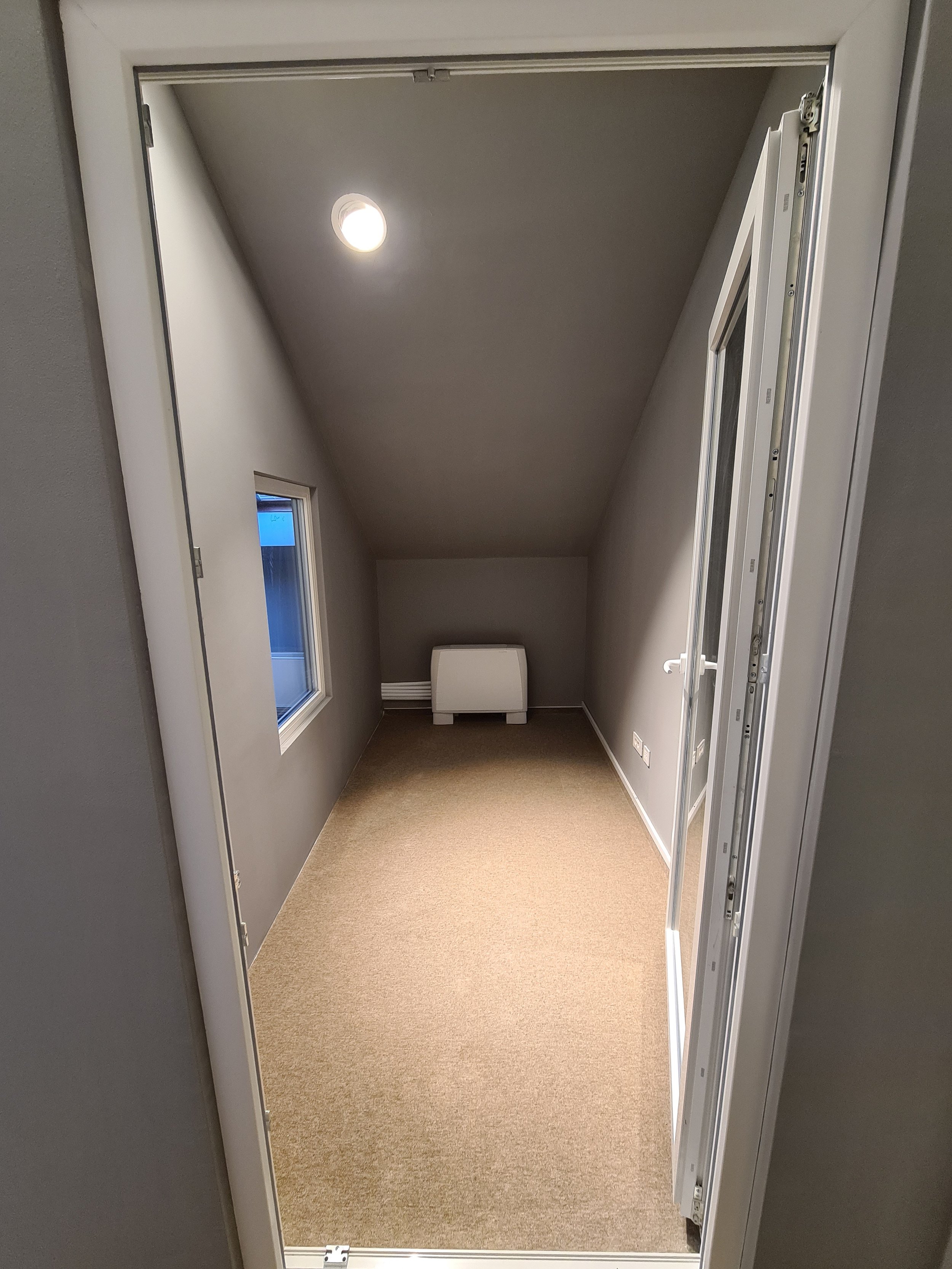 Small attic room with sloped ceiling, beige carpet, gray walls, a small window on the left wall, a ceiling light, and an electric heater at the far end.